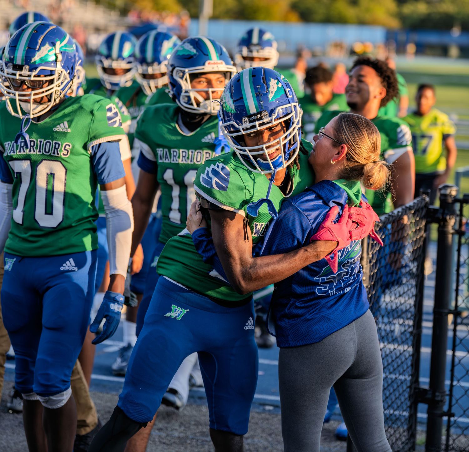 High school football player in green Warriors uniform receives hug from woman on sideline before game.