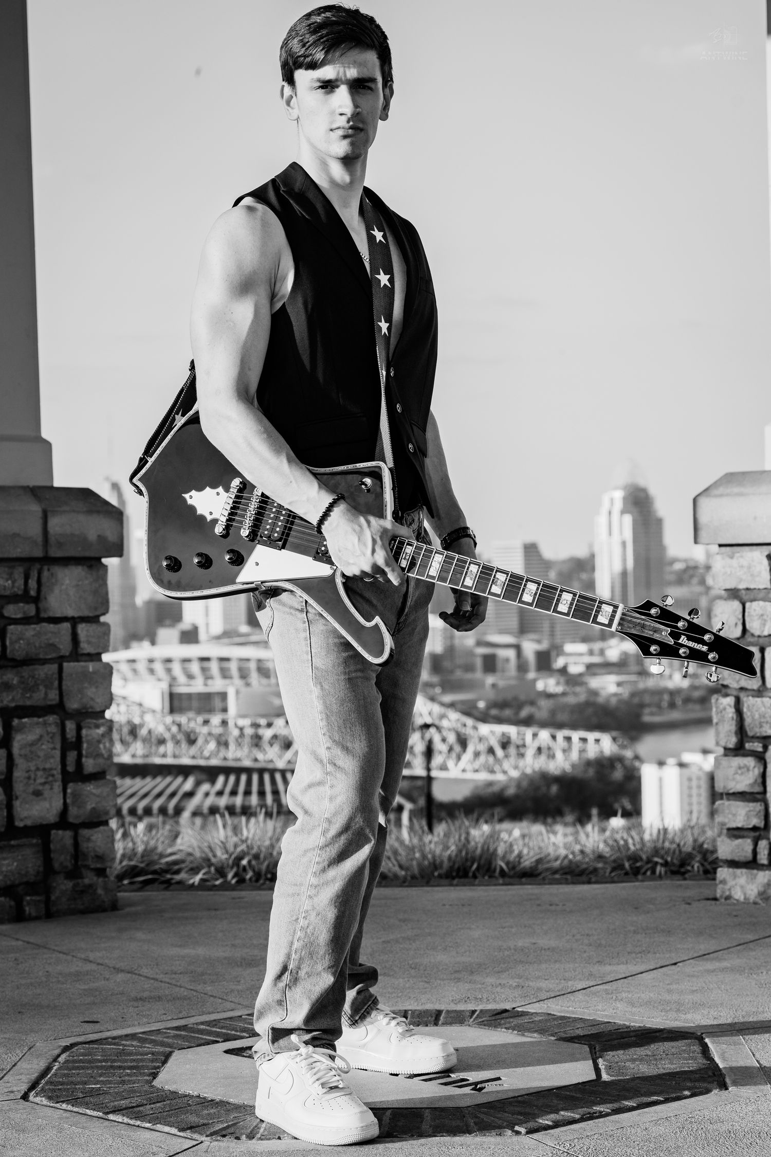 Young man in sleeveless vest holding electric guitar outdoors with city skyline in black and white photo.