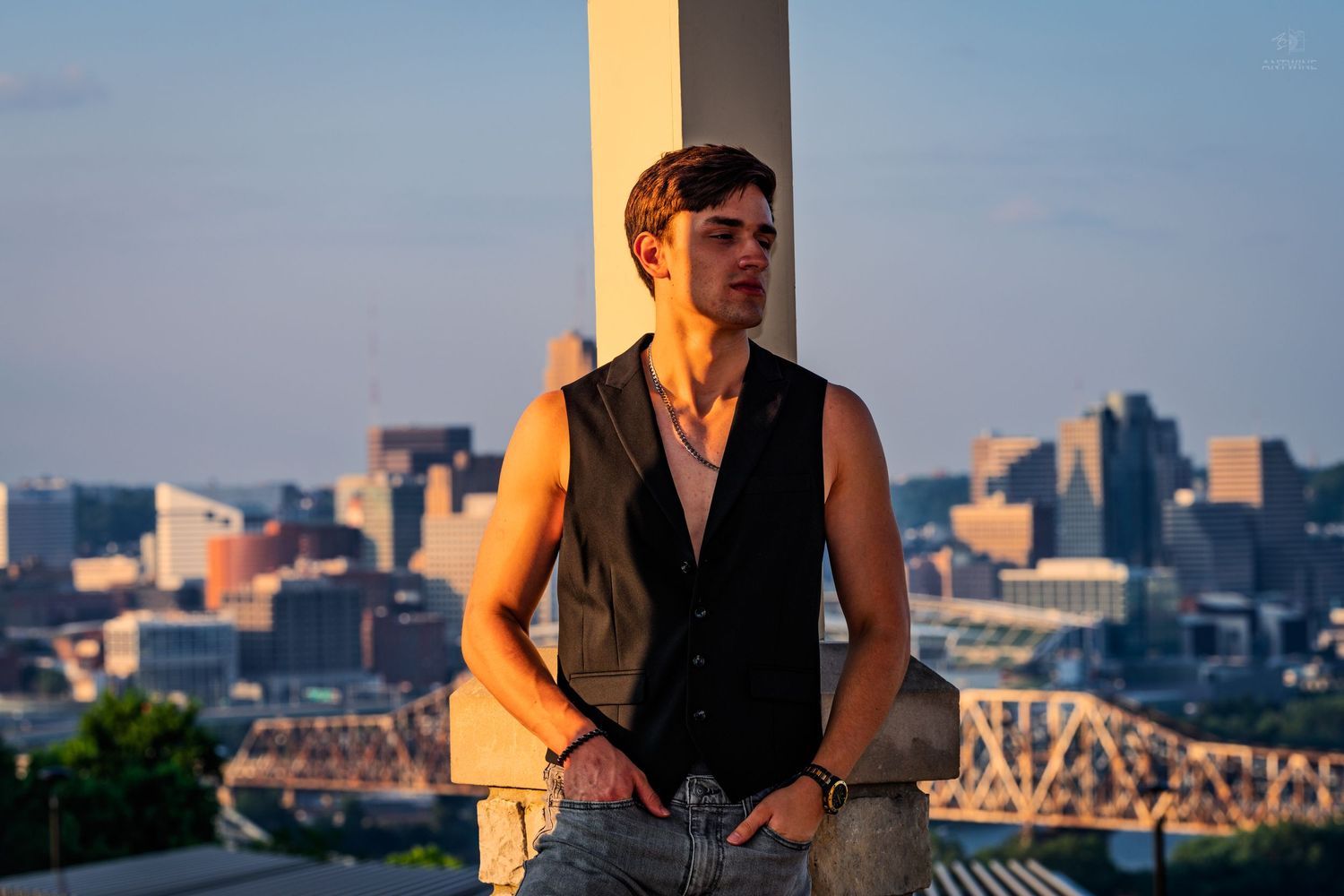 Young man in sleeveless vest poses against pillar with city skyline at golden hour.