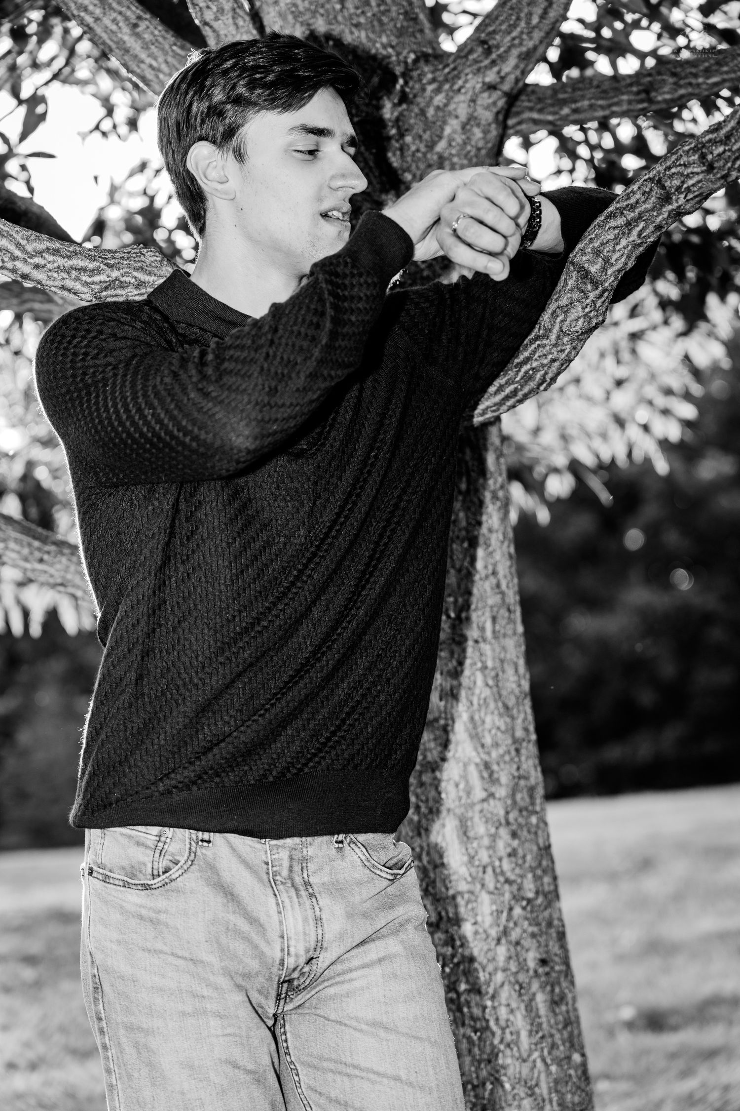 Black and white photo of a young man in a black sweater and jeans leaning against a tree outdoors.