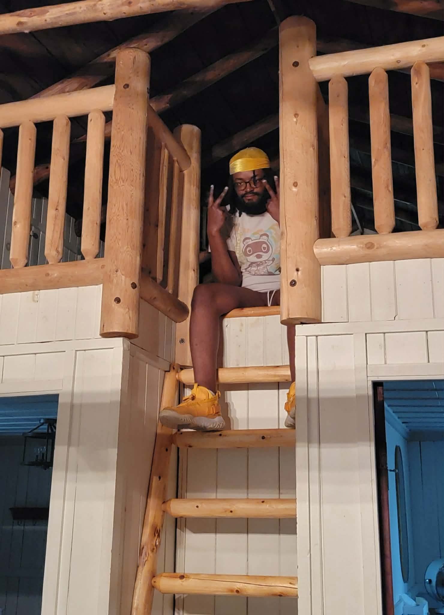 Man in yellow hat sitting on a wooden loft ladder inside a rustic log cabin interior.