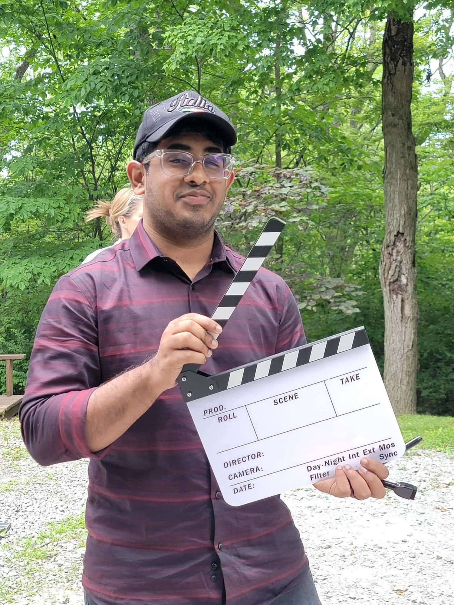 Man in striped shirt and cap holding a film clapperboard outdoors with green trees in background.