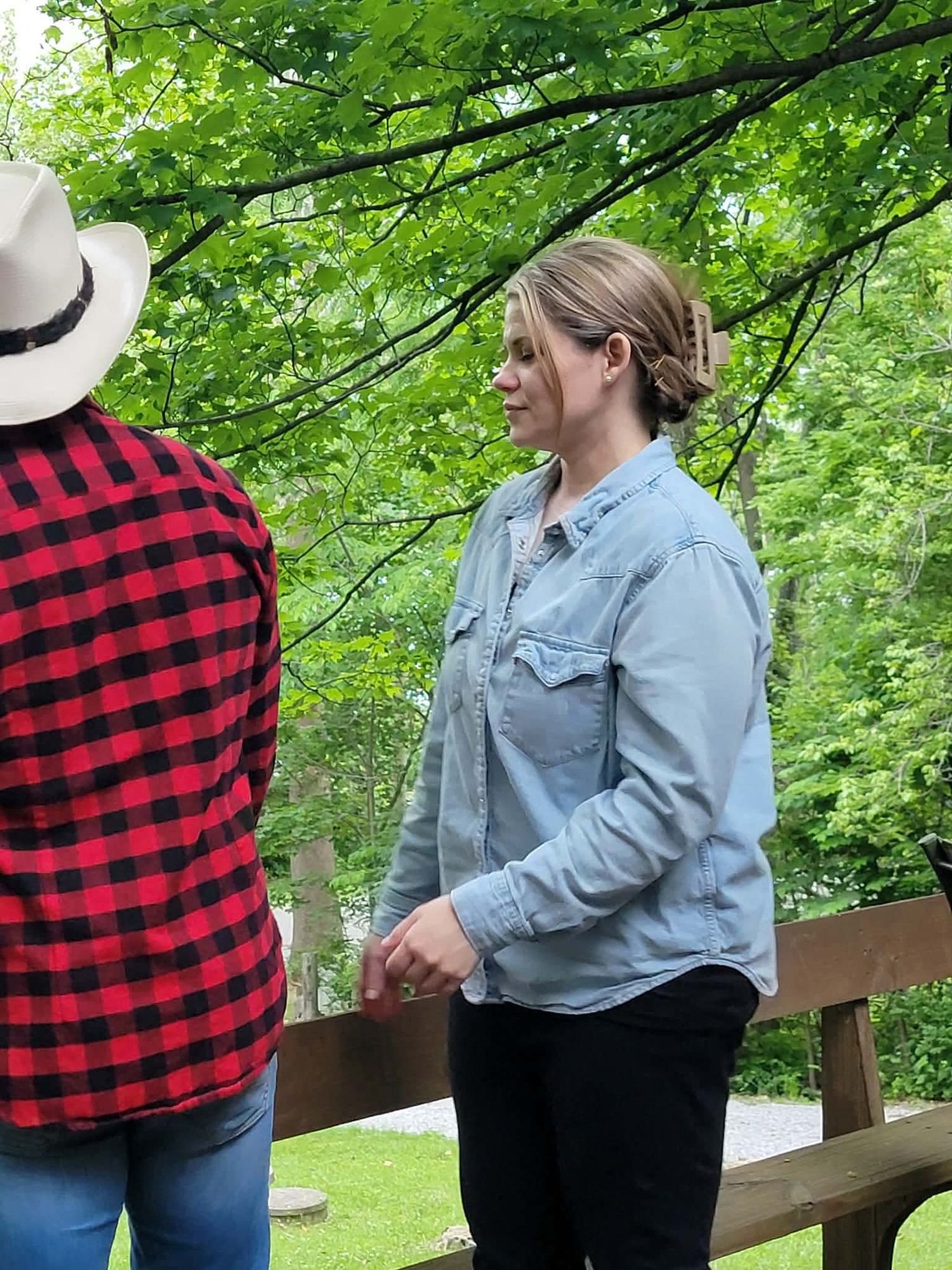 Woman in denim shirt stands on wooden deck conversing with person in red plaid shirt under green trees.
