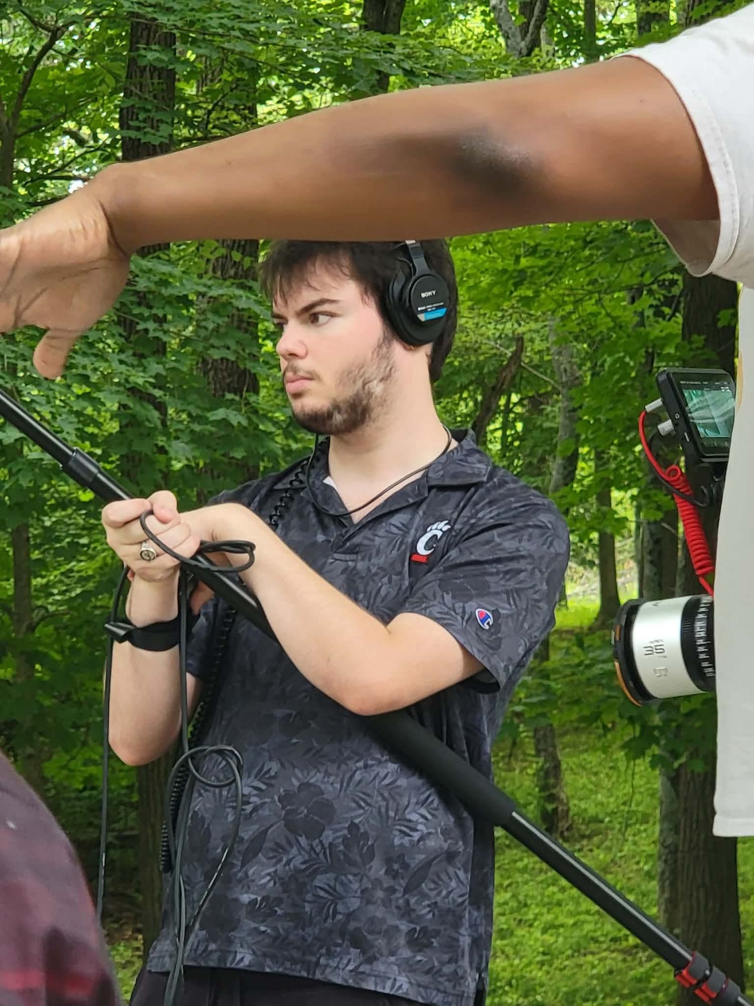 Young man with headphones holds a boom pole on an outdoor film set surrounded by green trees.