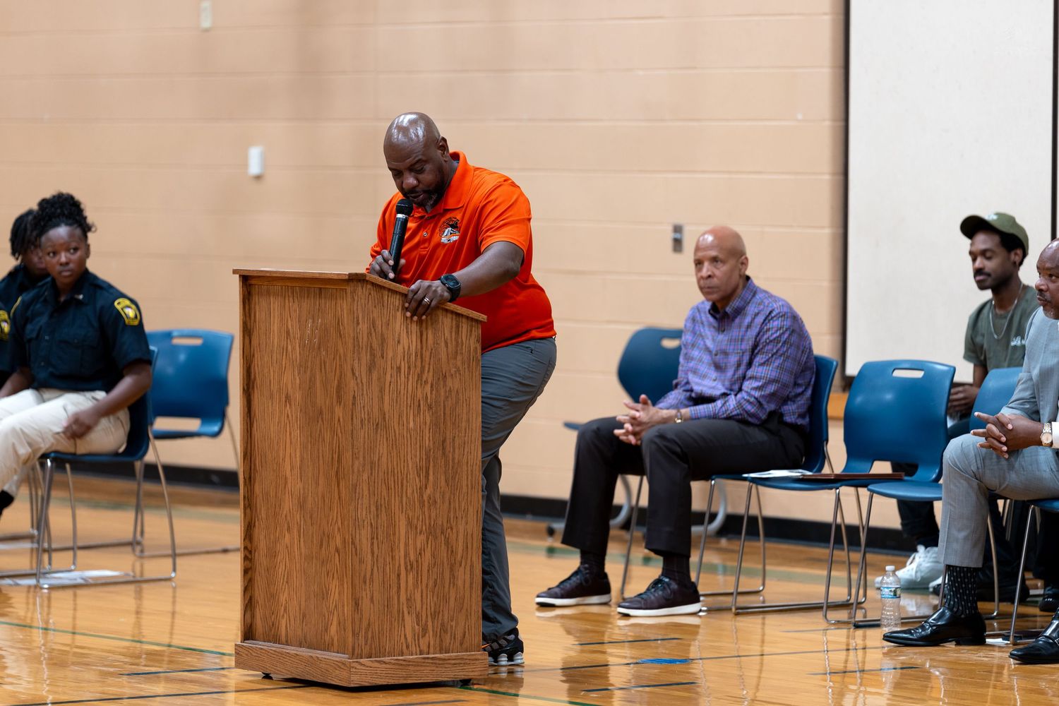 Man in orange shirt speaks at podium during community meeting in gymnasium with attendees seated in background.