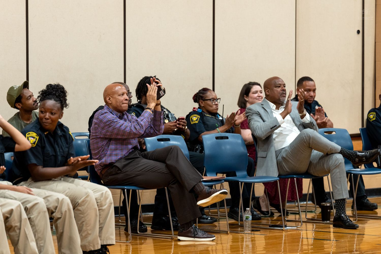 Community members and officials seated in a gymnasium during a meeting, with one person photographing the event.