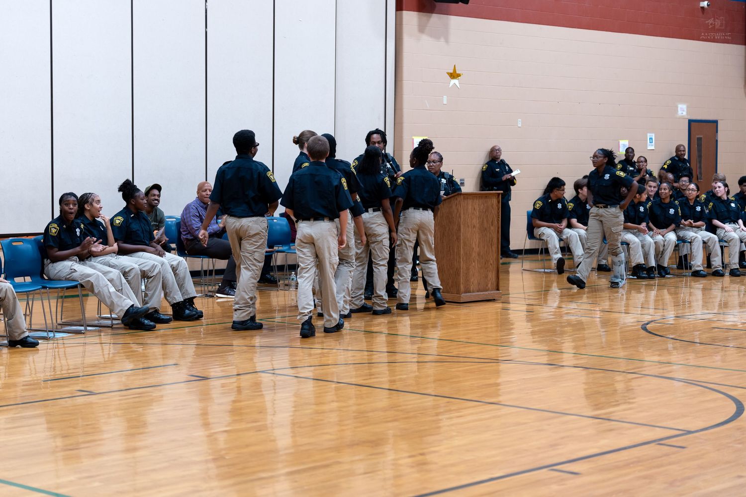Group of uniformed officers standing and sitting in a gymnasium during an indoor assembly or training event.