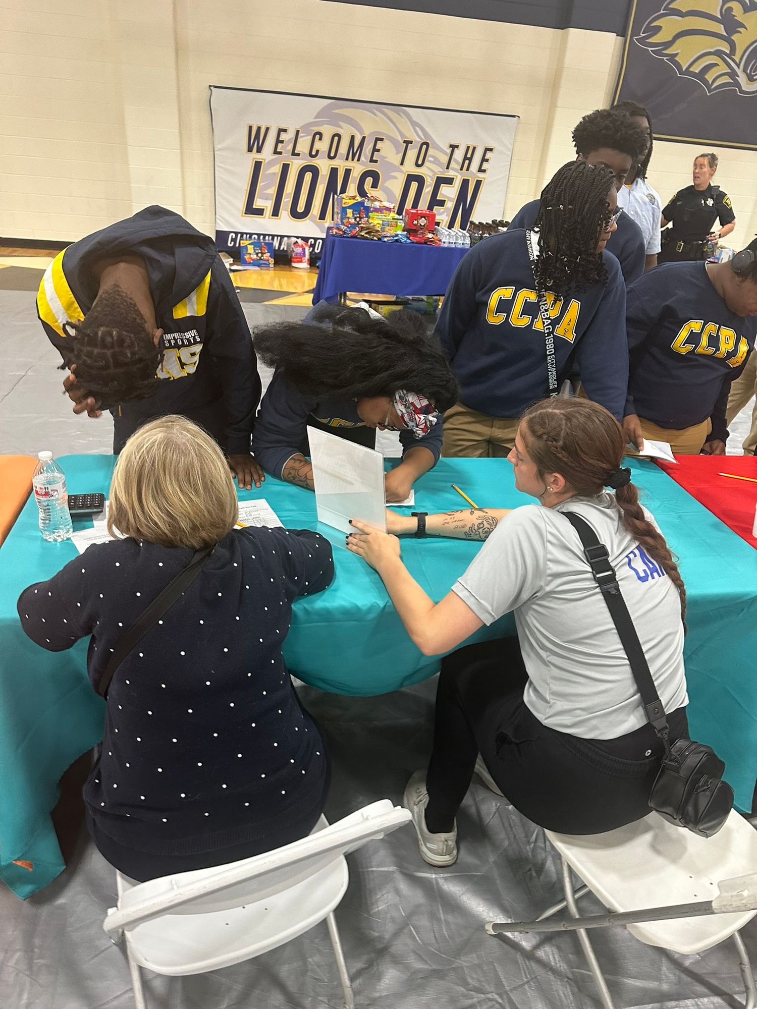 Students and adults collaborate at a teal table inside a school gymnasium during a Lions Den event.