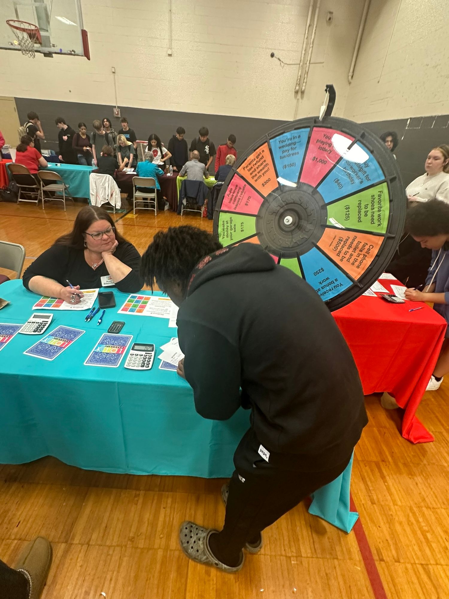 Students participate in a colorful prize wheel activity at a school gymnasium fair event with tables and attendees.