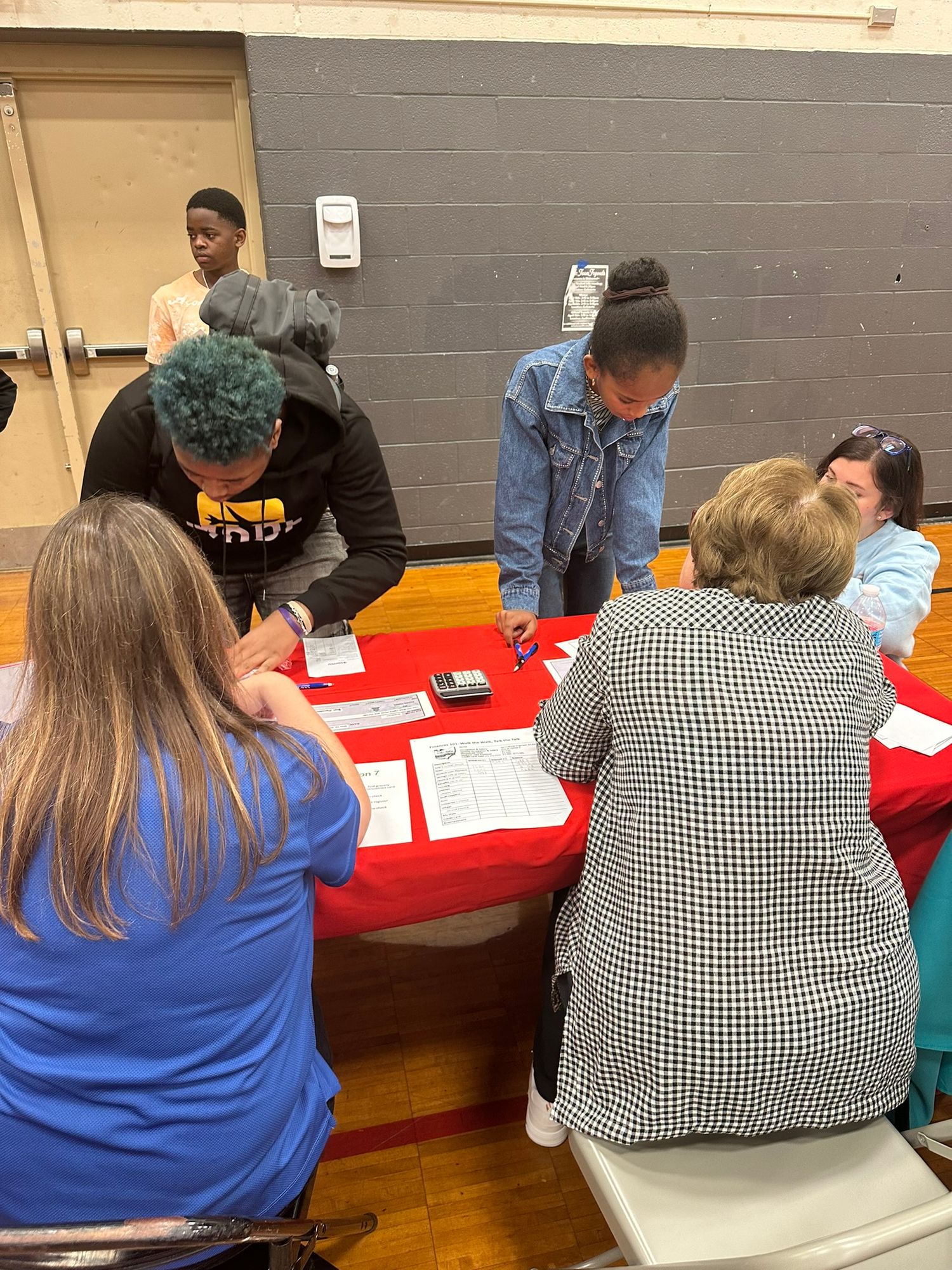 Students and adults collaborate around a red table in a school gymnasium during an educational event or registration fair.