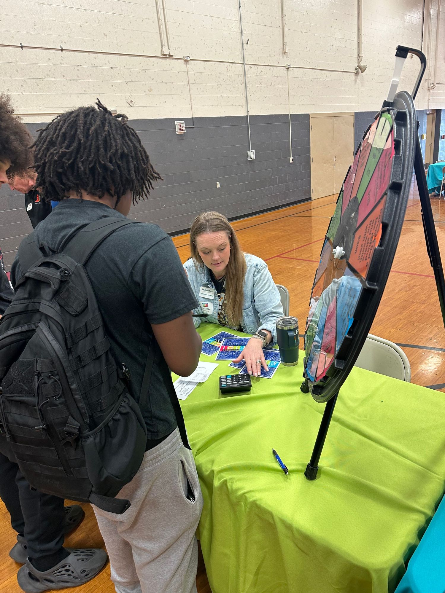 Students gather around a bright green table with a spin wheel at a school career fair event in a gymnasium.