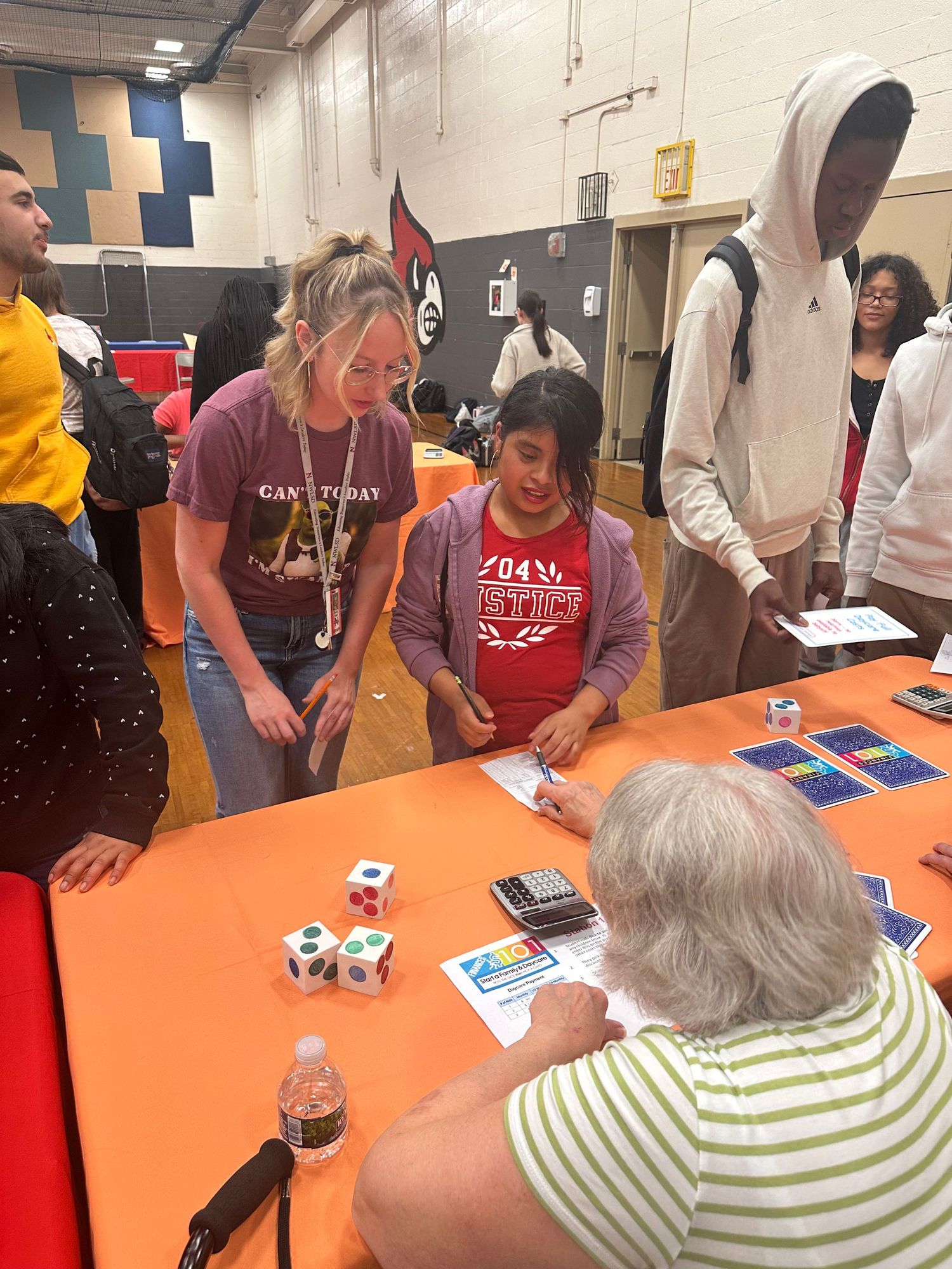 Students and teacher engage in a math dice game activity at an indoor school event with colorful tables.