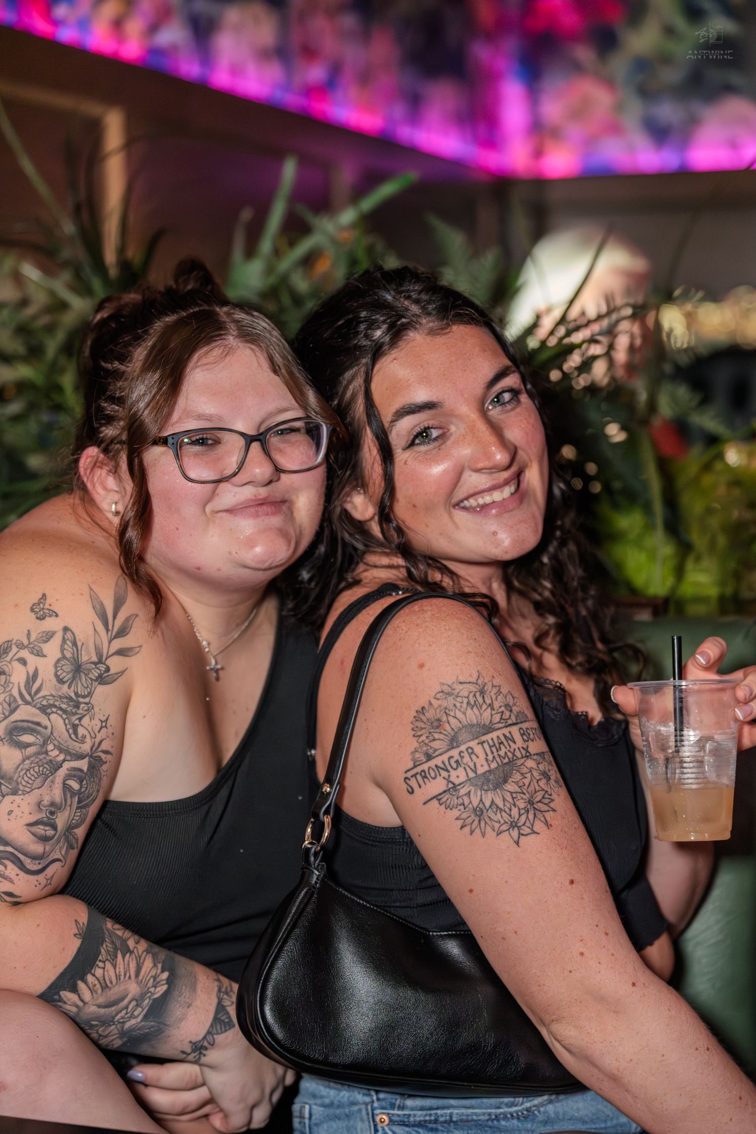Two women with tattoos in black tops pose together smiling at a plant-decorated nightclub lounge area.