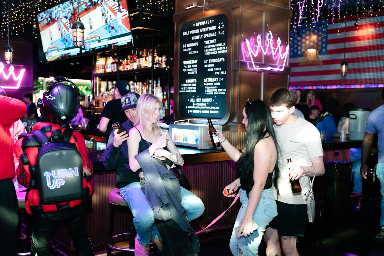 People socializing at a lively bar with neon signs, TVs, and VR gaming equipment.
