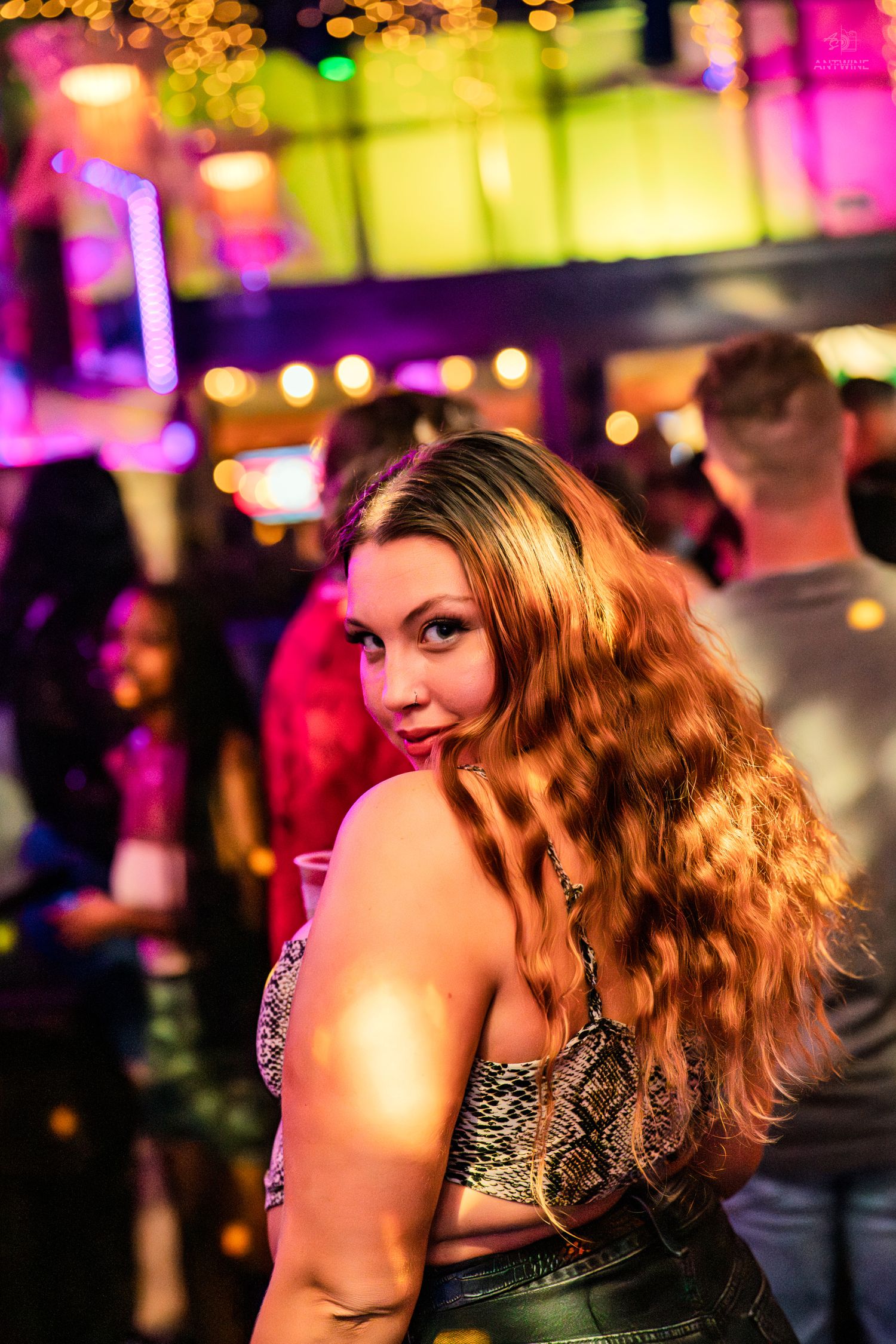 Woman with long curly auburn hair posing at a colorful nightclub with bokeh lights in background.