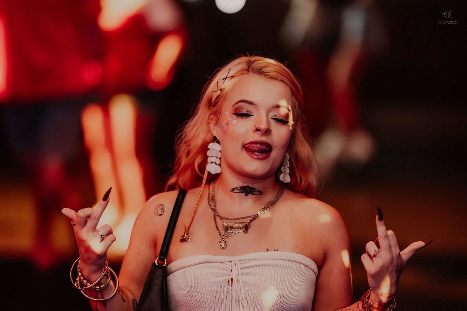 Blonde woman with glitter makeup and tattoos poses expressively at a nightclub event.