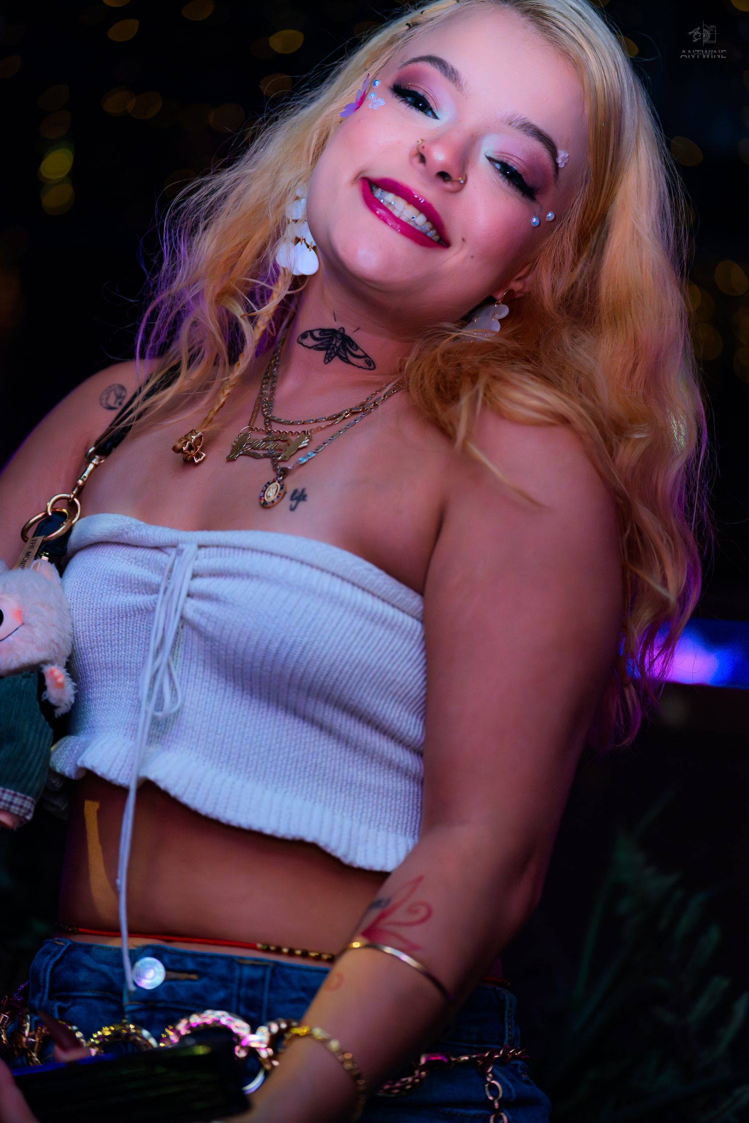 Smiling blonde woman in white tube top and denim shorts with festival makeup and layered necklaces at night.