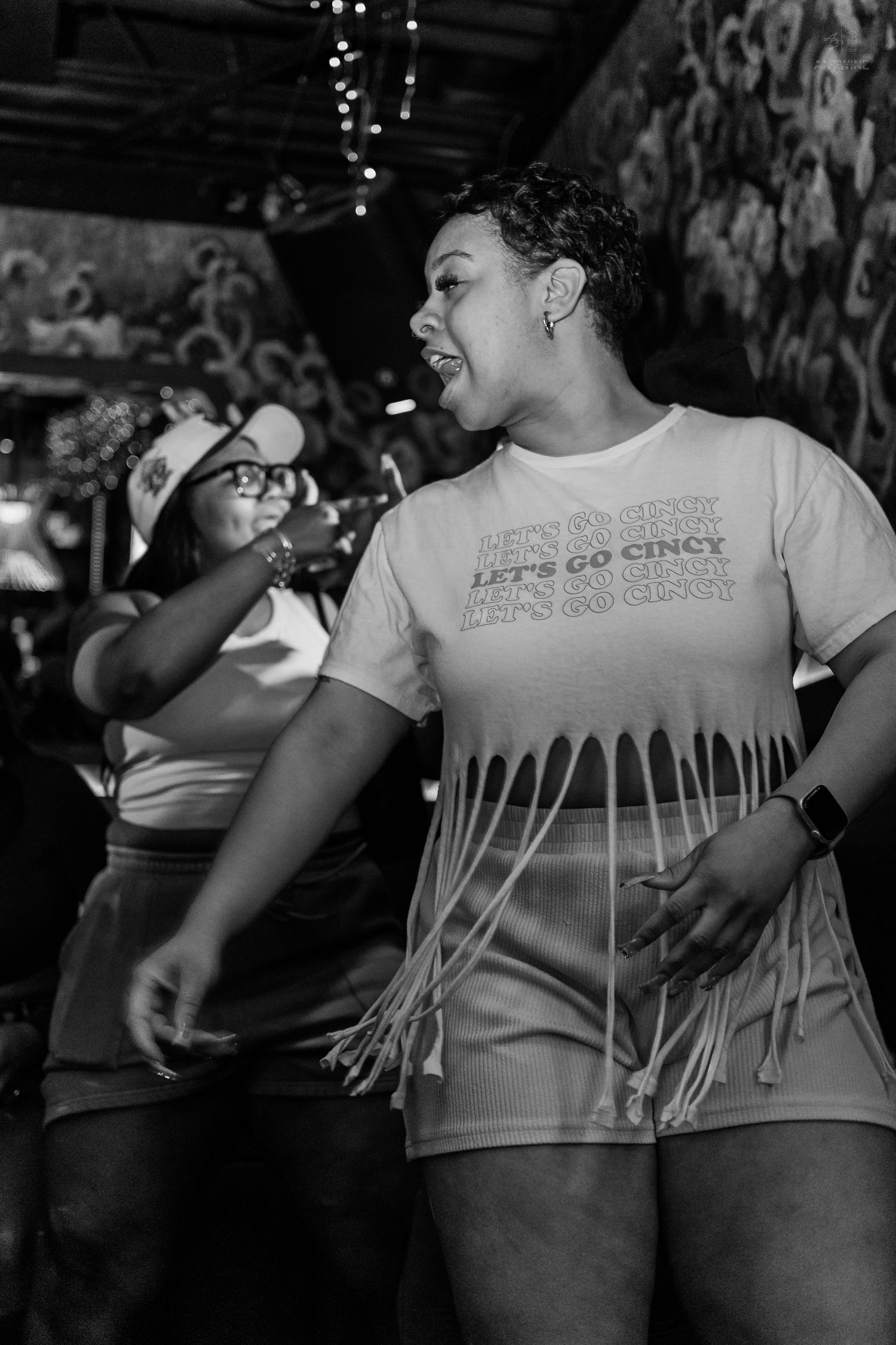 Woman dancing joyfully in a Let's Go Cincy fringe shirt at a lively bar event in black and white.