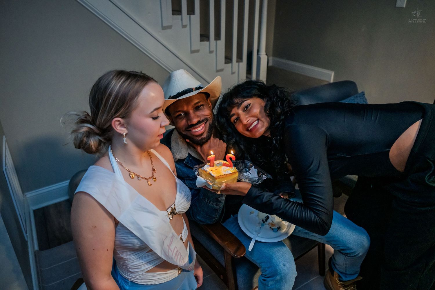 Three friends gathered around a small birthday cake with lit candles on a staircase at a party.