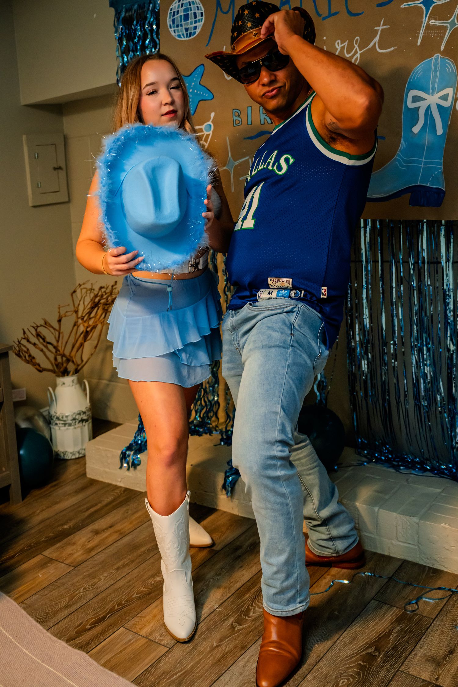 Woman holding blue feathered cowboy hat poses with man in Dallas Mavericks jersey at party.