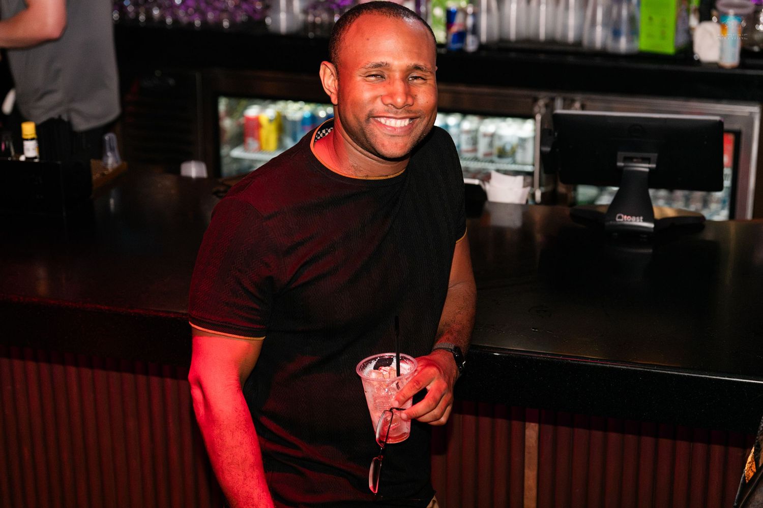 Smiling man in black t-shirt holds a pink cocktail at a bar.