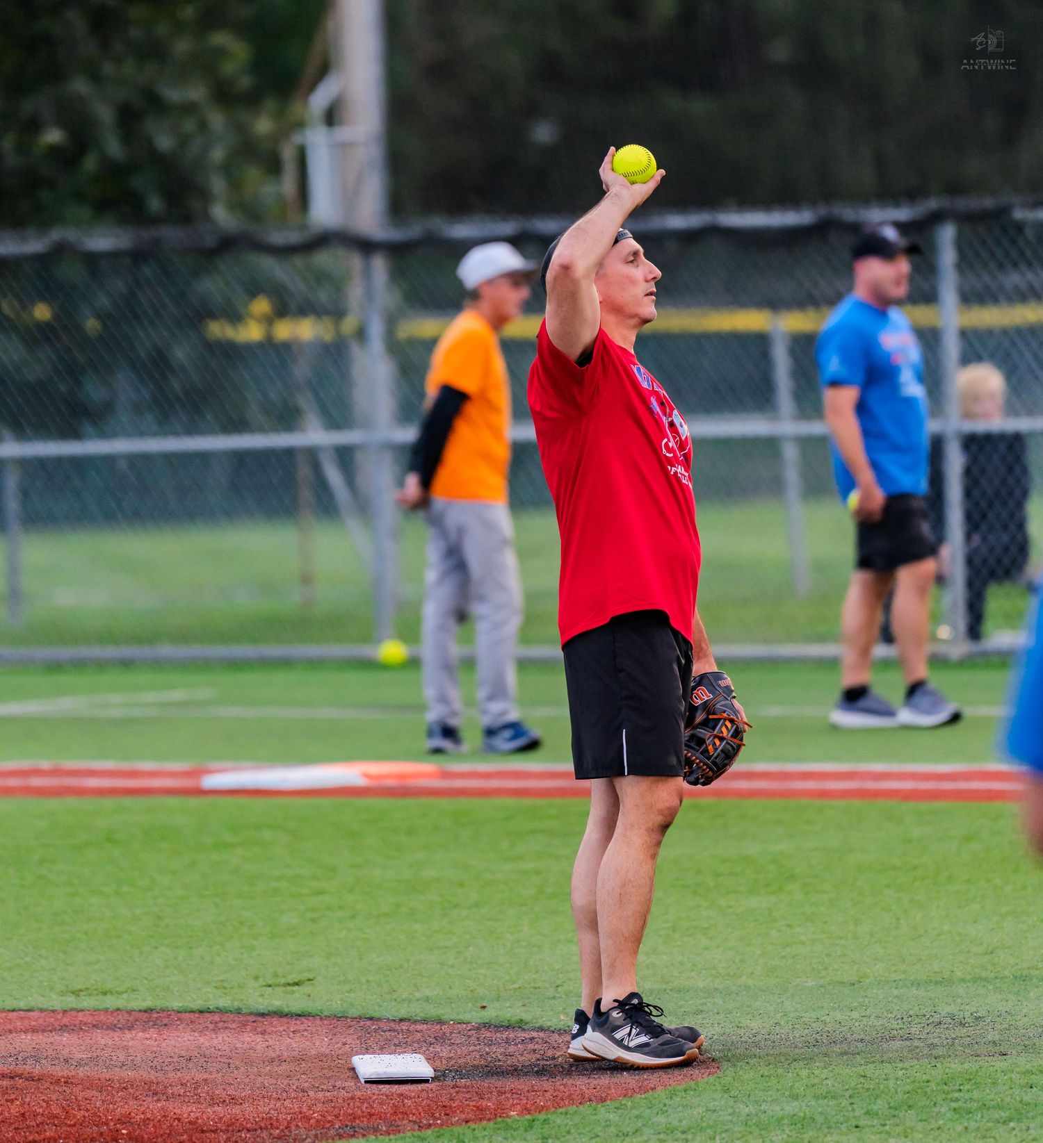A softball pitcher in a red shirt winds up to throw a yellow ball on a dirt mound during an outdoor recreational game.