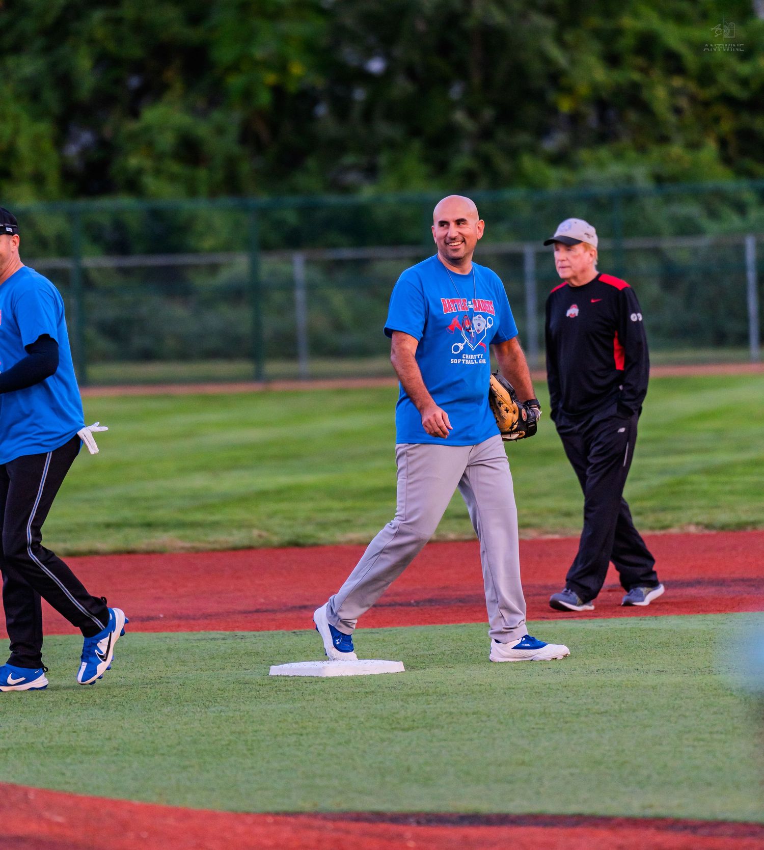 Three men in blue and red athletic wear stand on a baseball diamond during an outdoor charity softball game.
