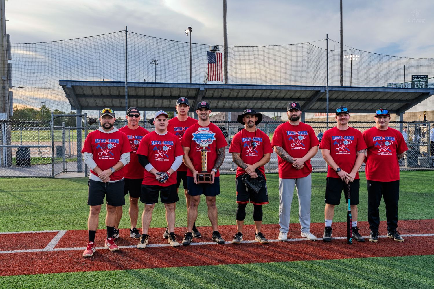 A softball team in matching red shirts poses together on a baseball field holding a championship plaque.