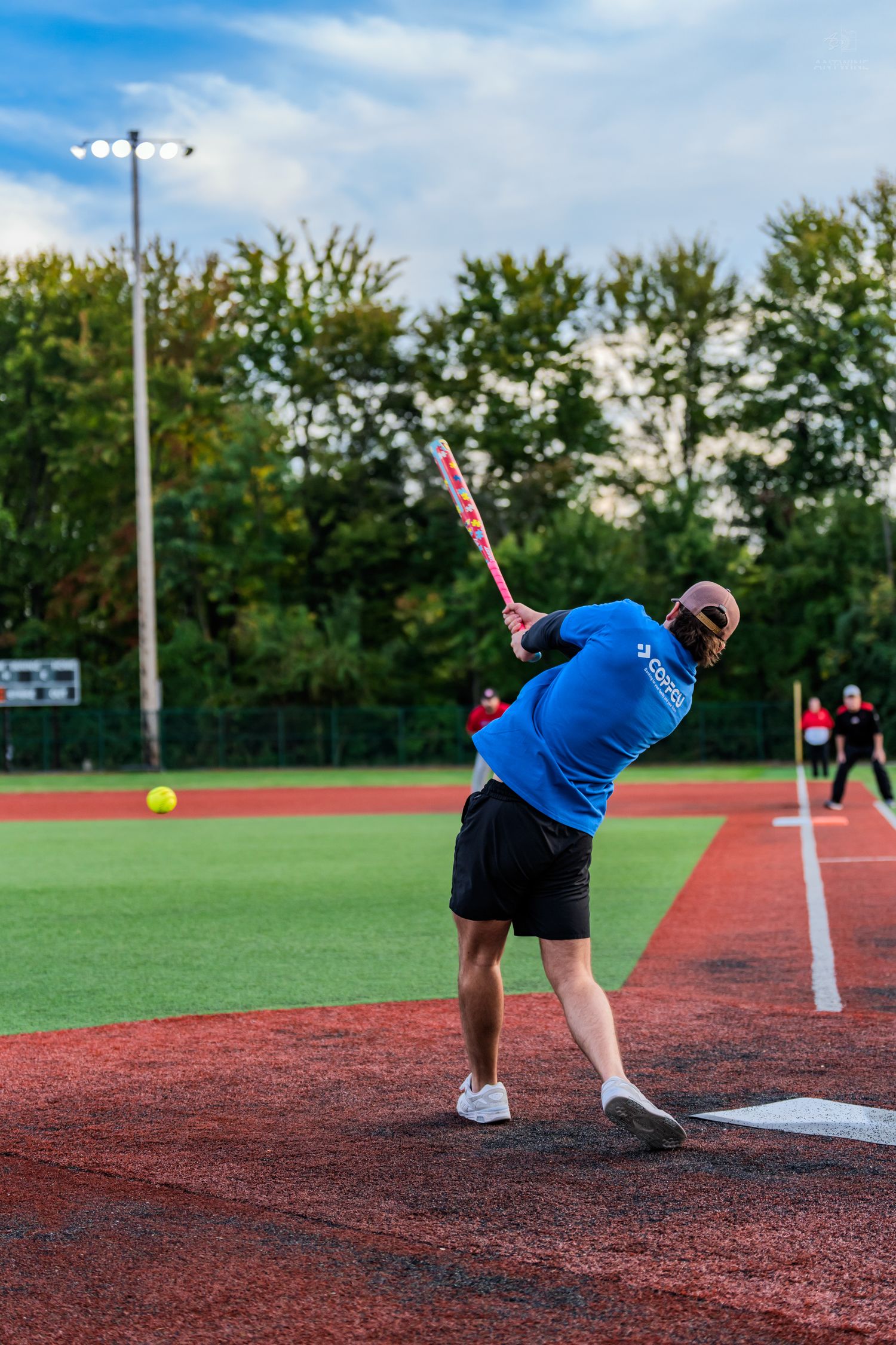 A batter in a blue shirt swings at a yellow softball on a red turf field during an outdoor recreational game.
