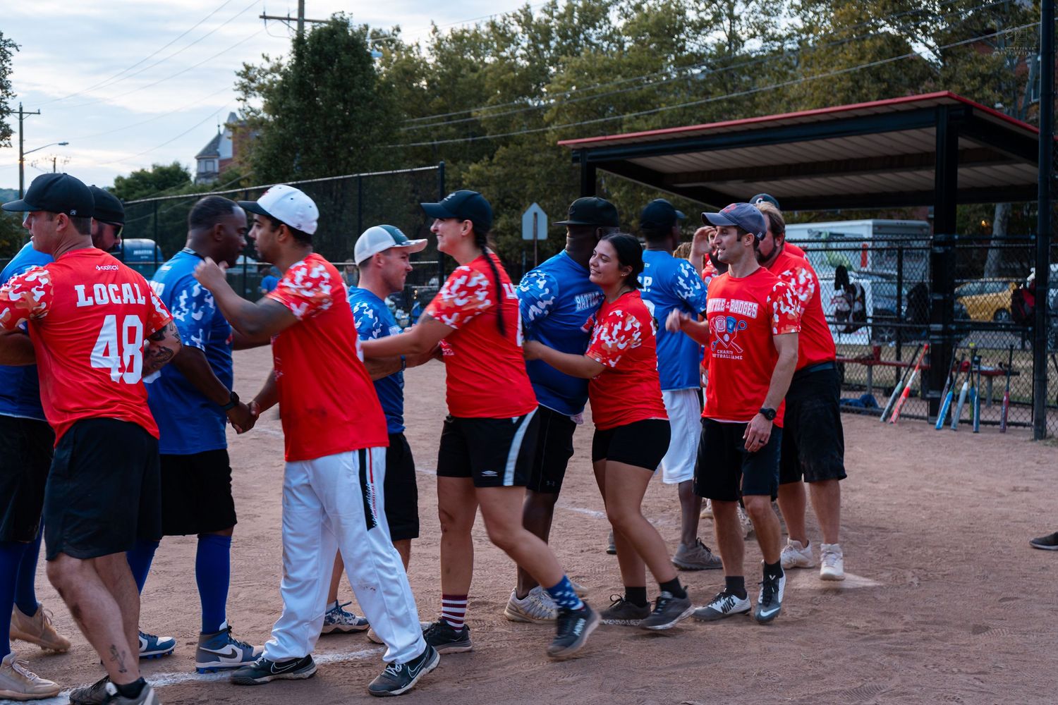 Baseball players in red and blue uniforms shaking hands after a game at an outdoor softball field.