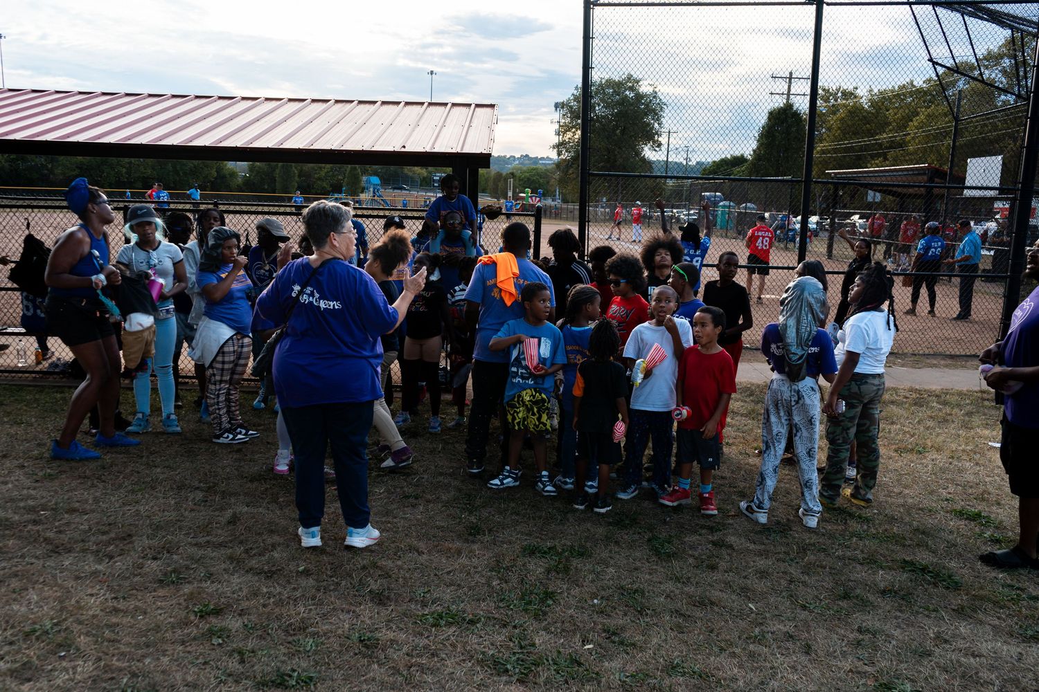 Children and adults gather at a softball field during an outdoor youth sports event at dusk.