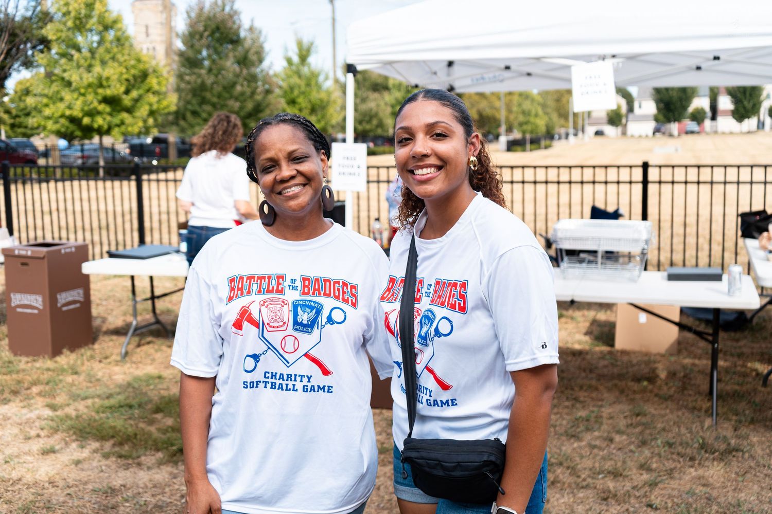 Two women smile wearing Battle of the Brews charity softball game t-shirts outdoors.