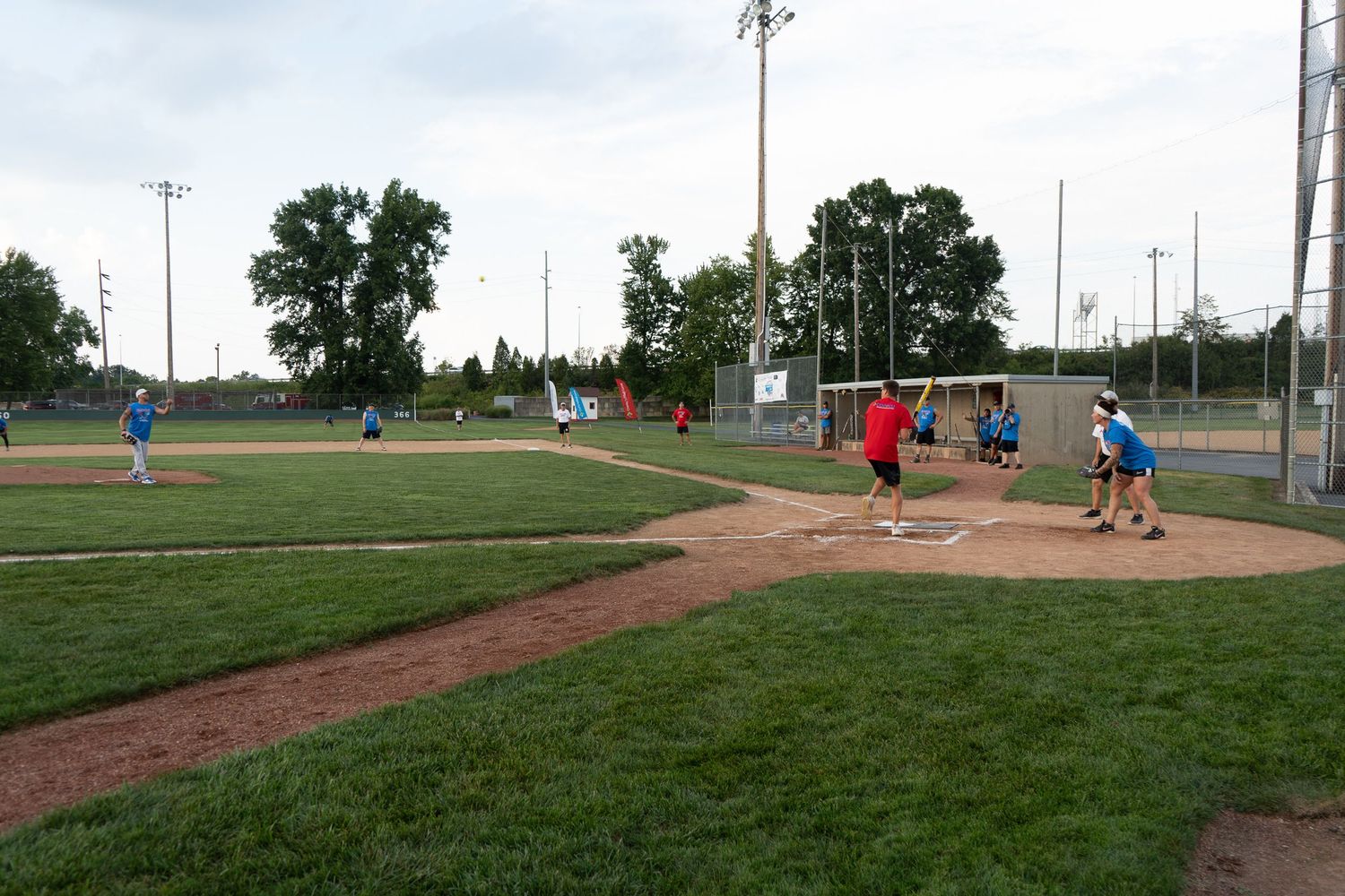 Teams compete in a recreational softball game on a green baseball field during a summer evening.
