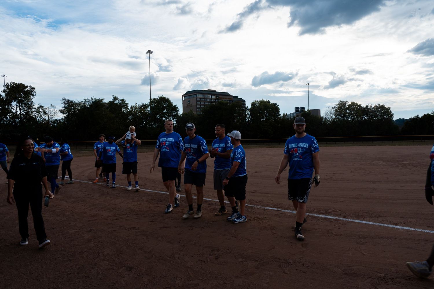 A team in blue shirts walks across a dirt baseball field at dusk with trees and a building in the background.