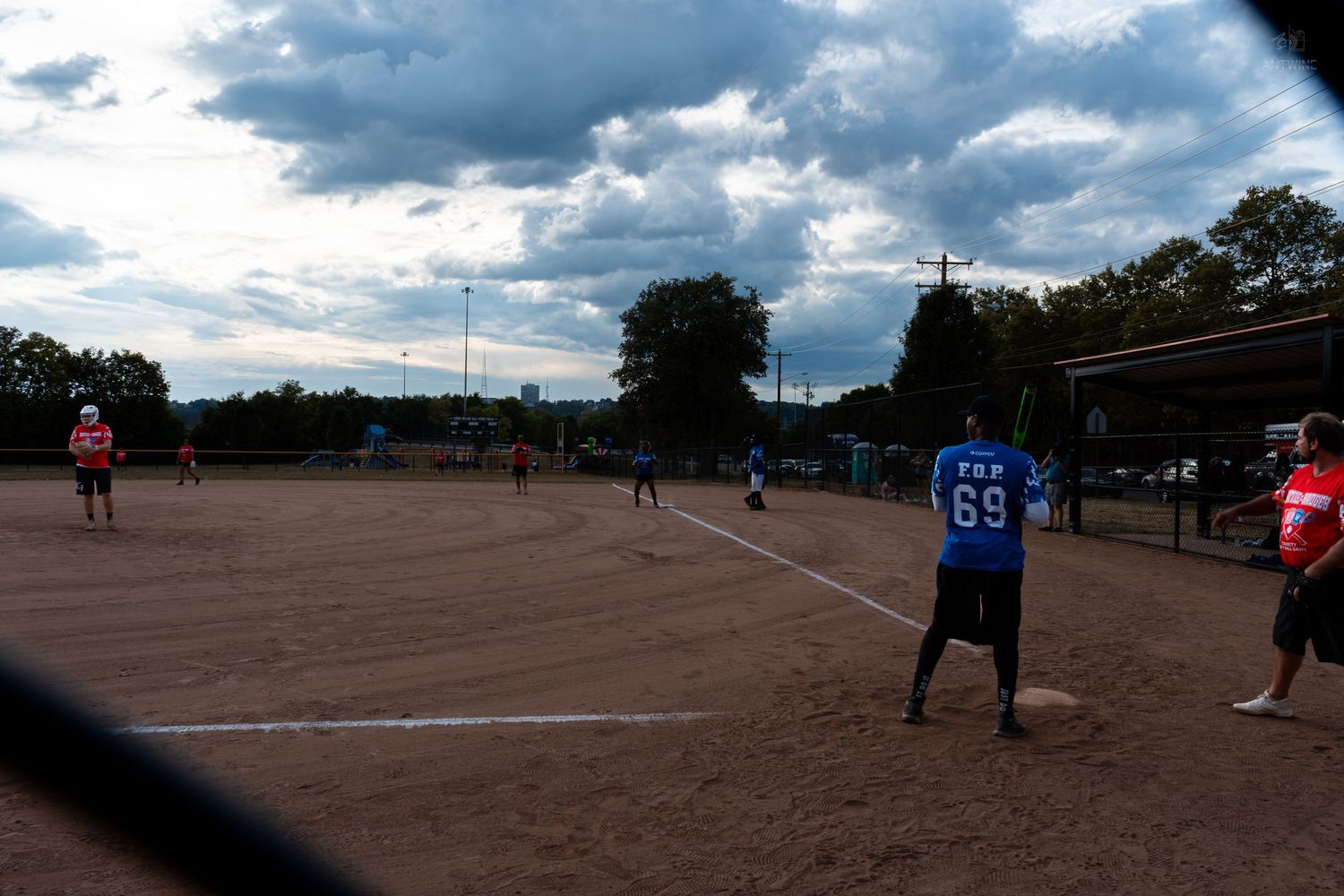 Softball players on a dirt field under cloudy skies, with player number 69 in blue ready to field the ball.