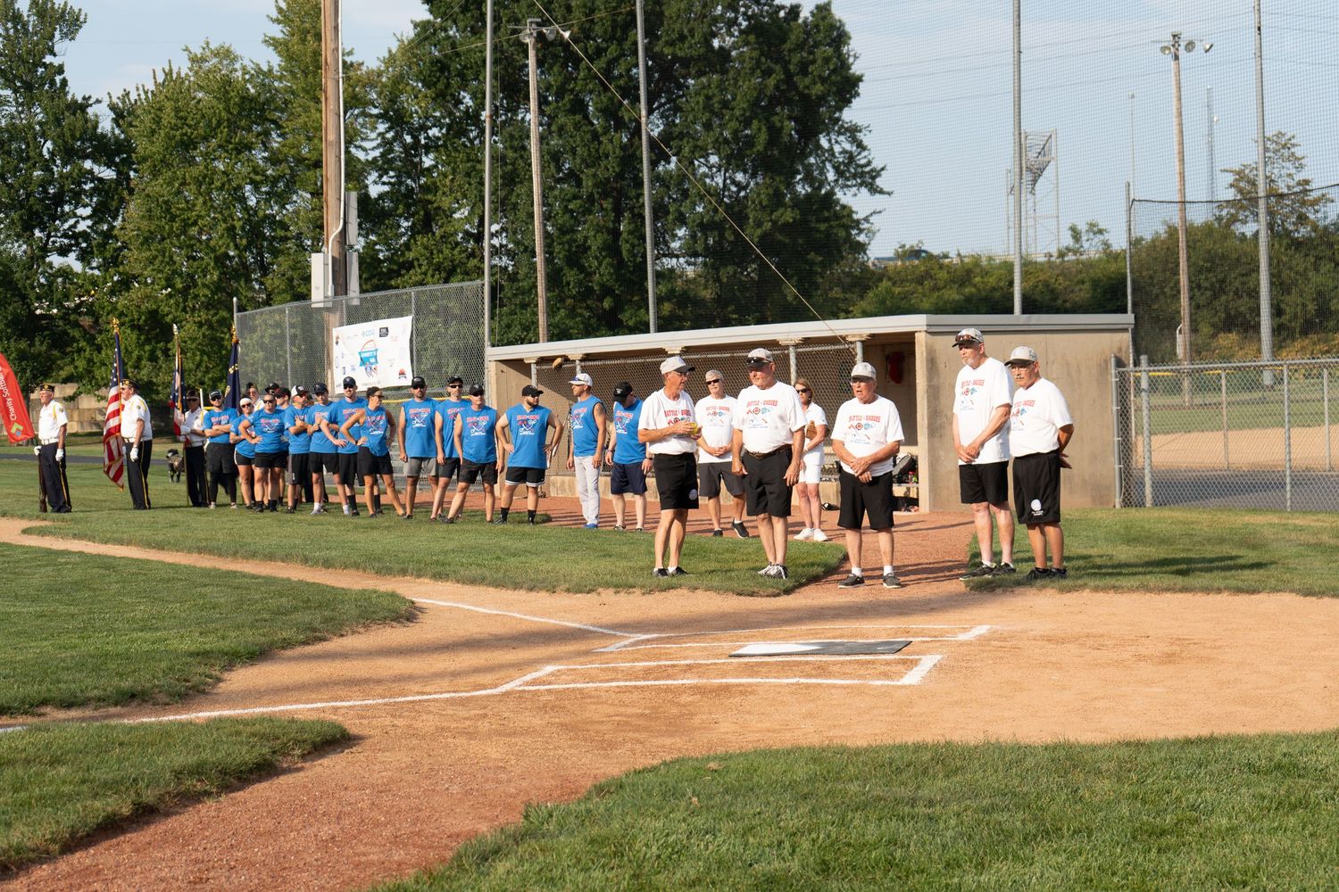 Teams line up on a baseball field during a ceremonial opening event with an American flag visible nearby.