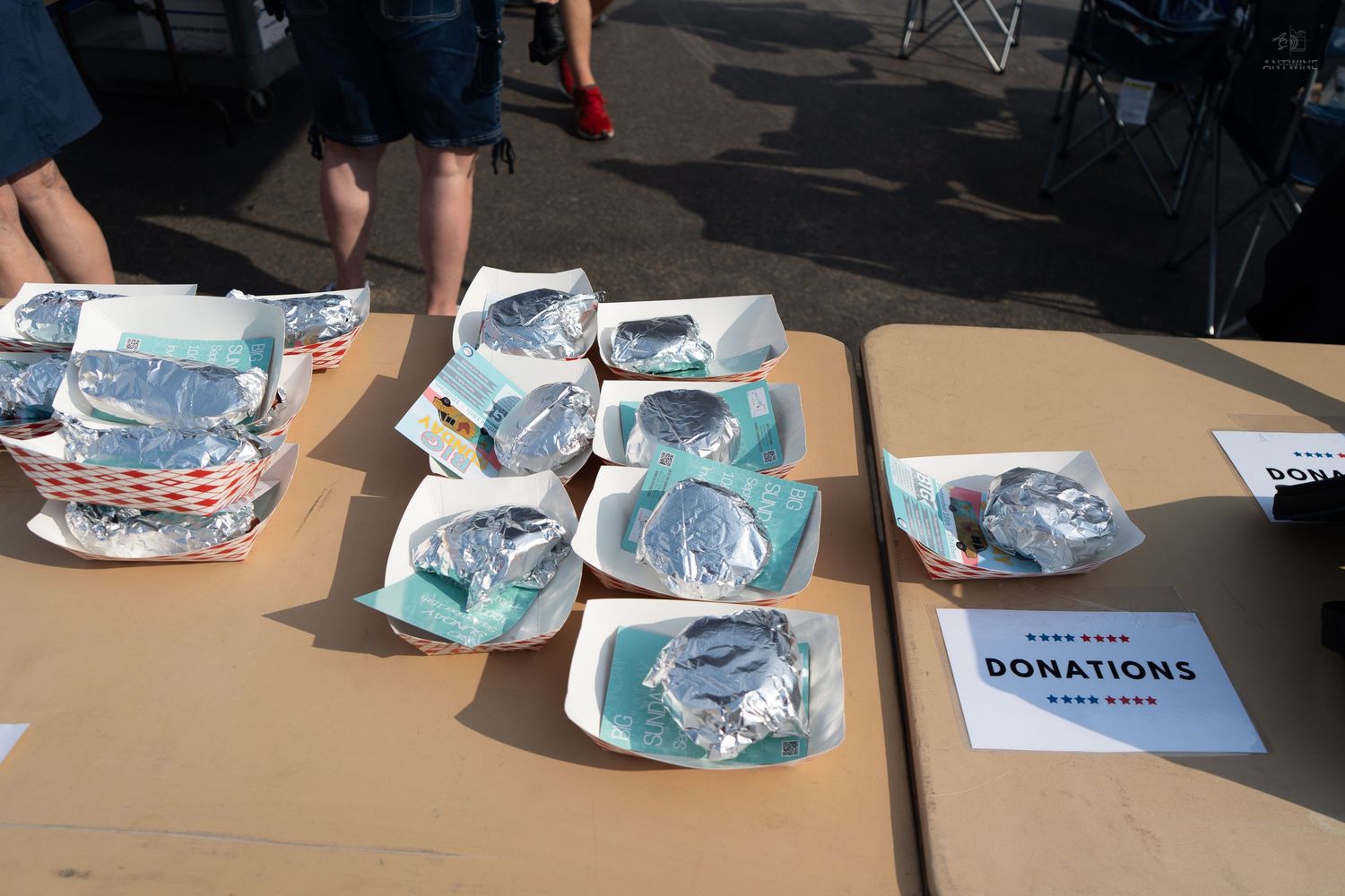 Outdoor event table with foil-covered food trays and a donations sign on a cardboard table.