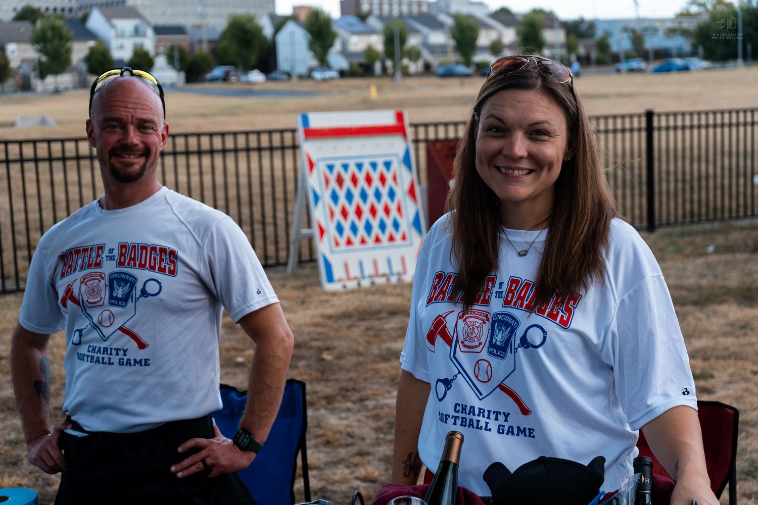 Two people smiling in matching charity softball game t-shirts at an outdoor event.