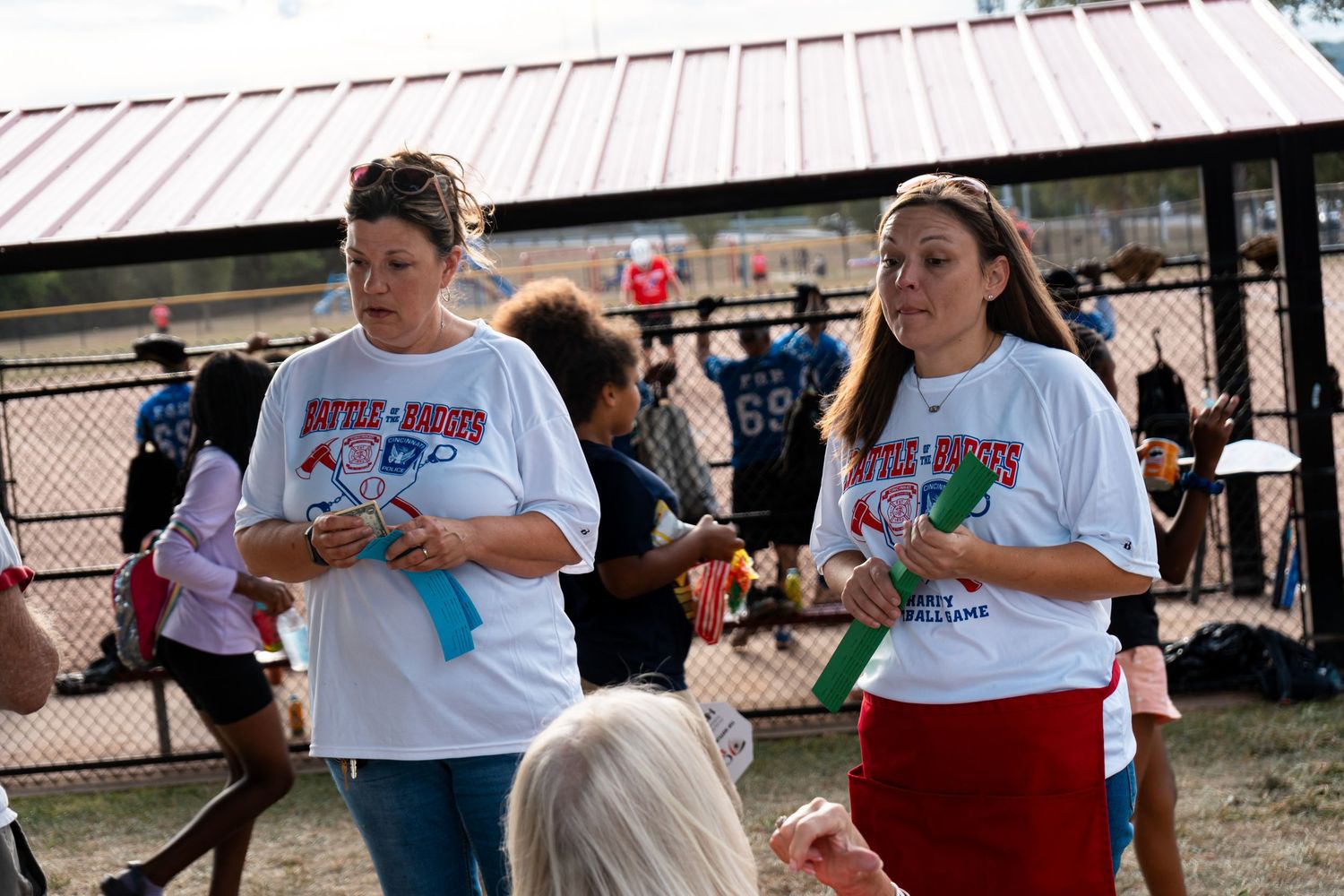 Two women in matching white Battle of the Badges event t-shirts stand outdoors at a community softball game.