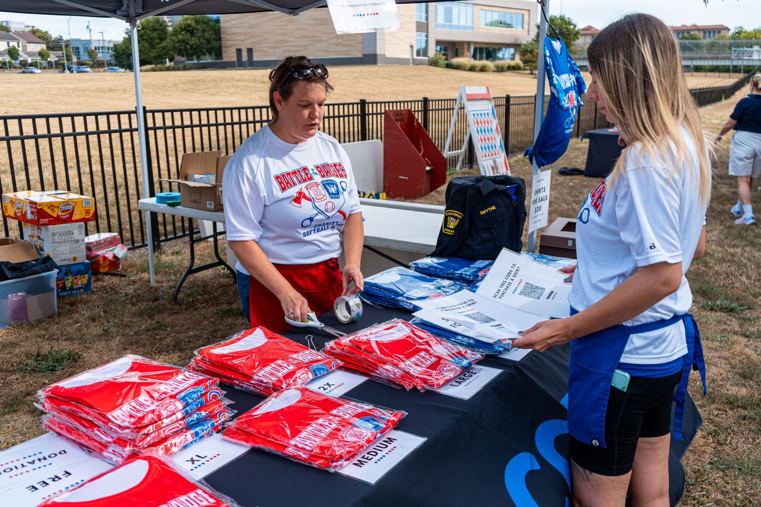 Two women distribute red and blue T-shirts at an outdoor event registration table under a tent on a sunny day.