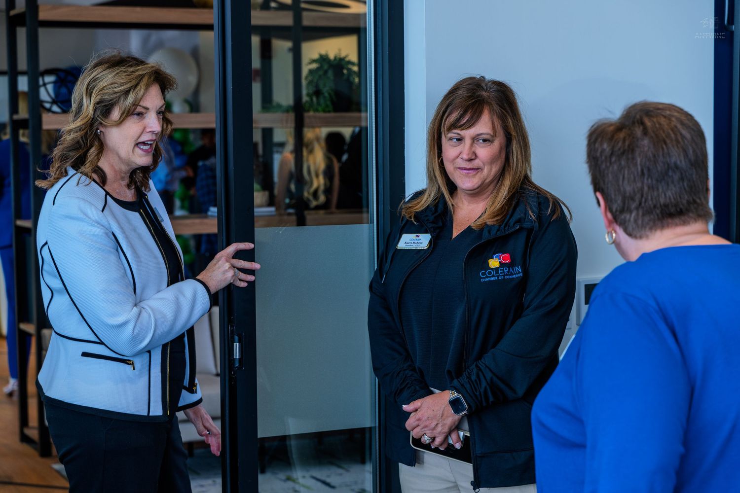 Three professional women engaged in conversation near a glass door in a modern office setting.