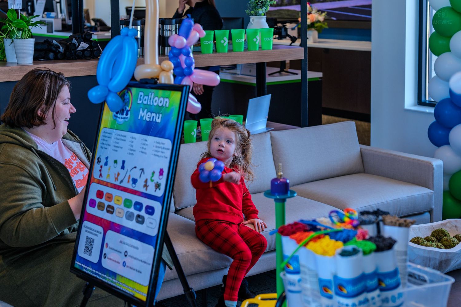Young child in red outfit sits near a balloon menu sign at a COPFCU grand opening event.