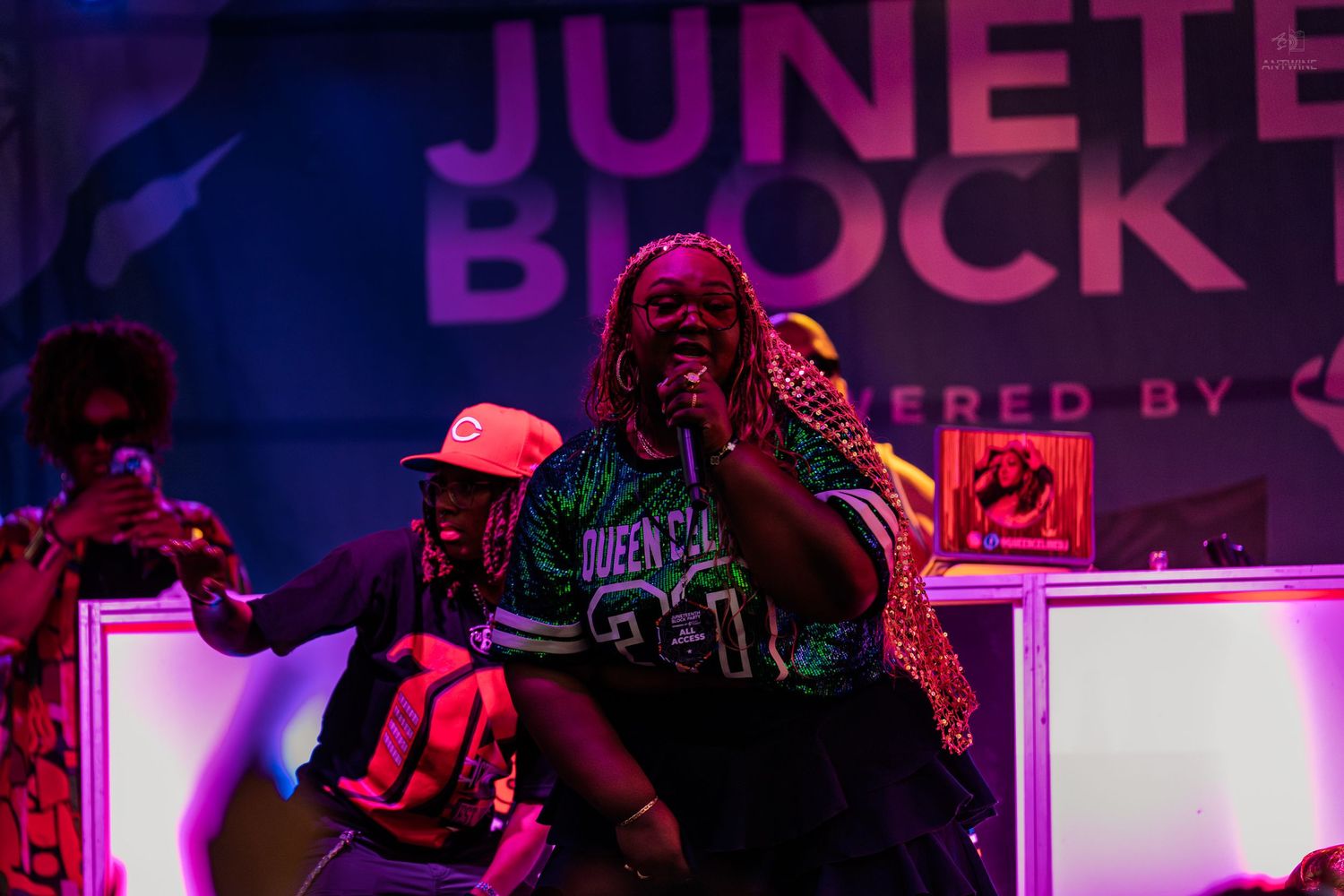 Female performer singing into microphone on stage at annual Juneteenth Block Party under purple lights.