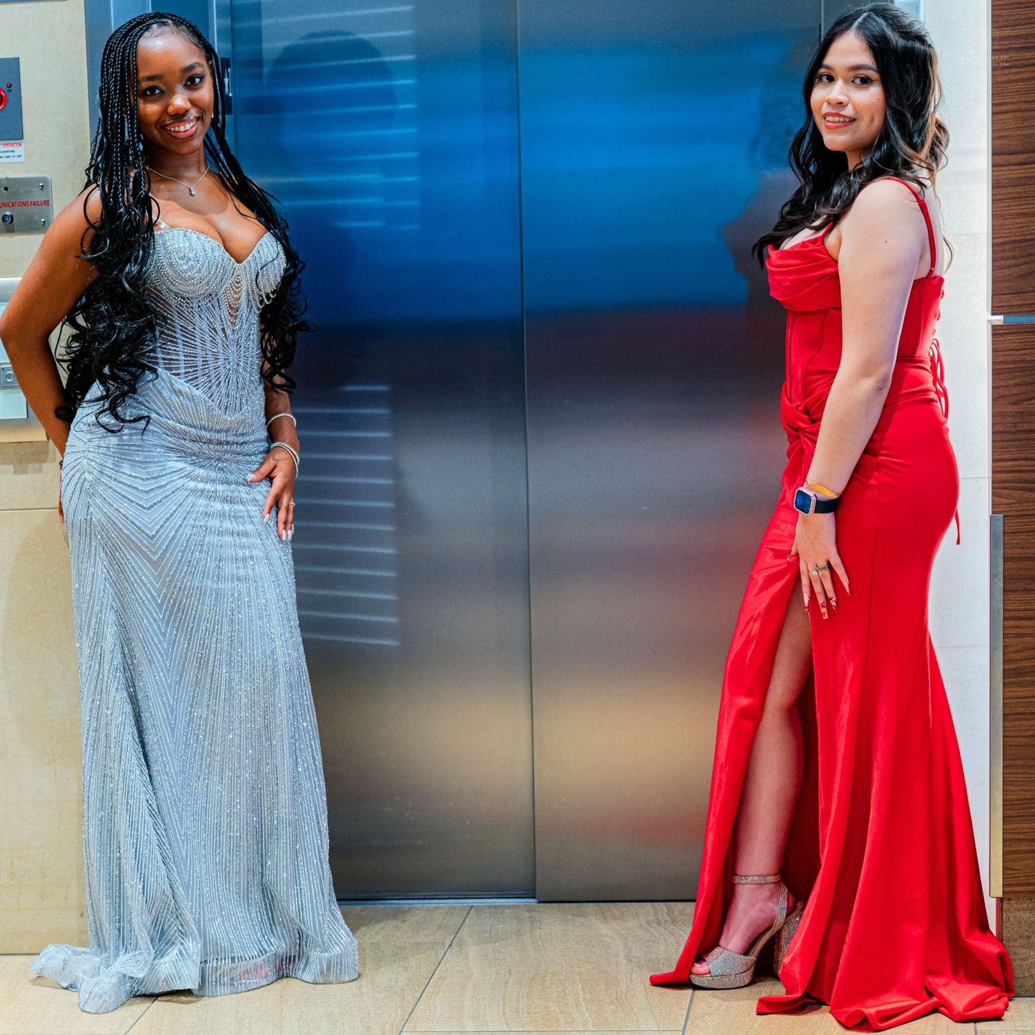 Two women in elegant prom gowns, one silver and one red mermaid dress, posing in front of an elevator.