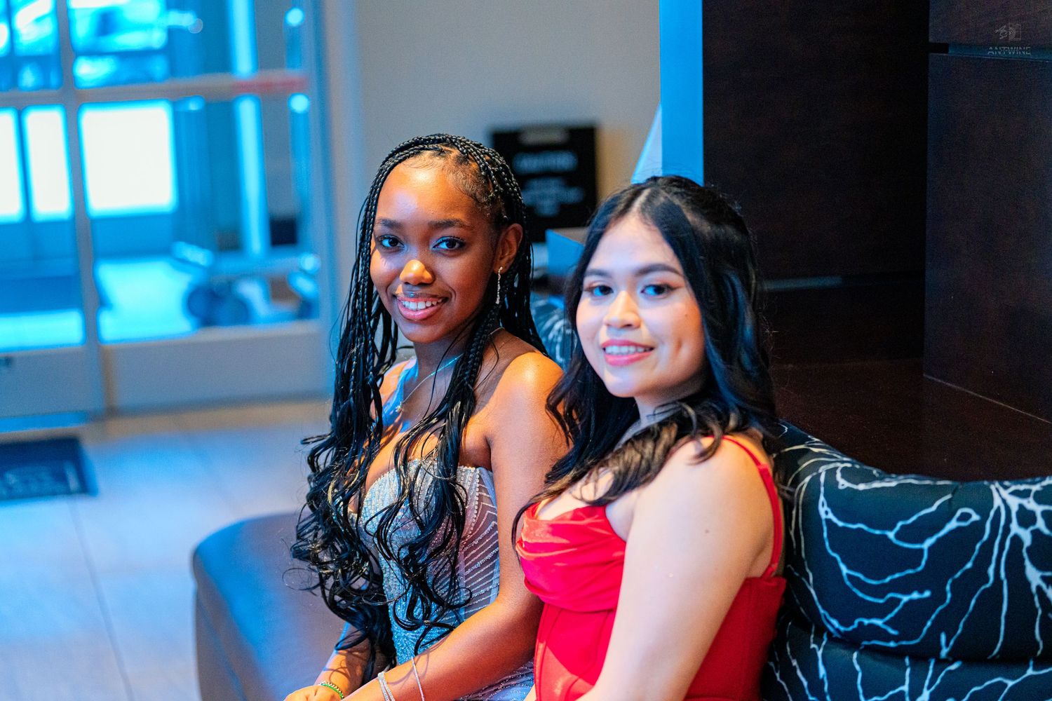 Two smiling young women in formal attire sitting closely together on a sofa for a portrait photo.