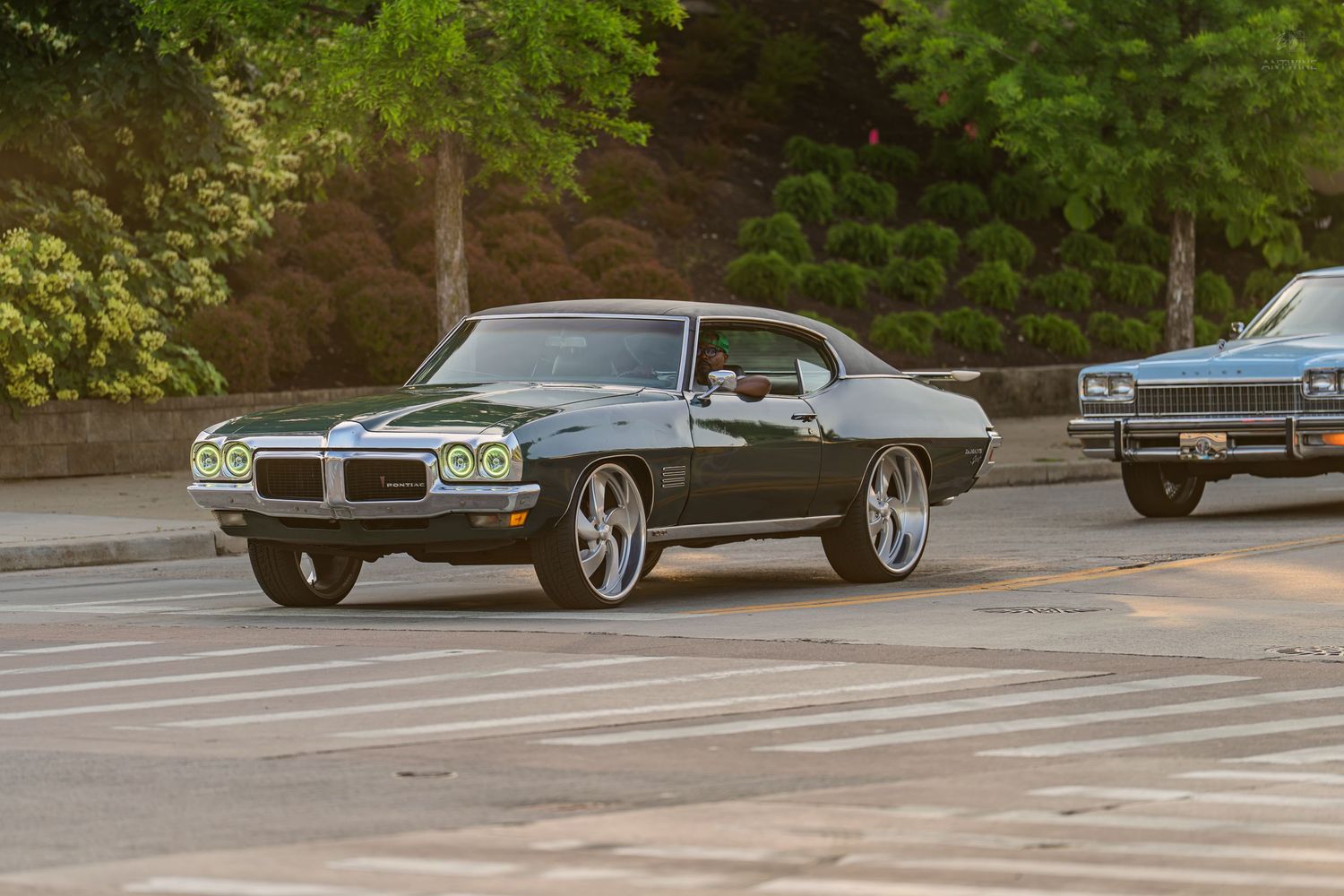Classic green Pontiac muscle car cruising on a sunny street with chrome wheels and vintage styling.