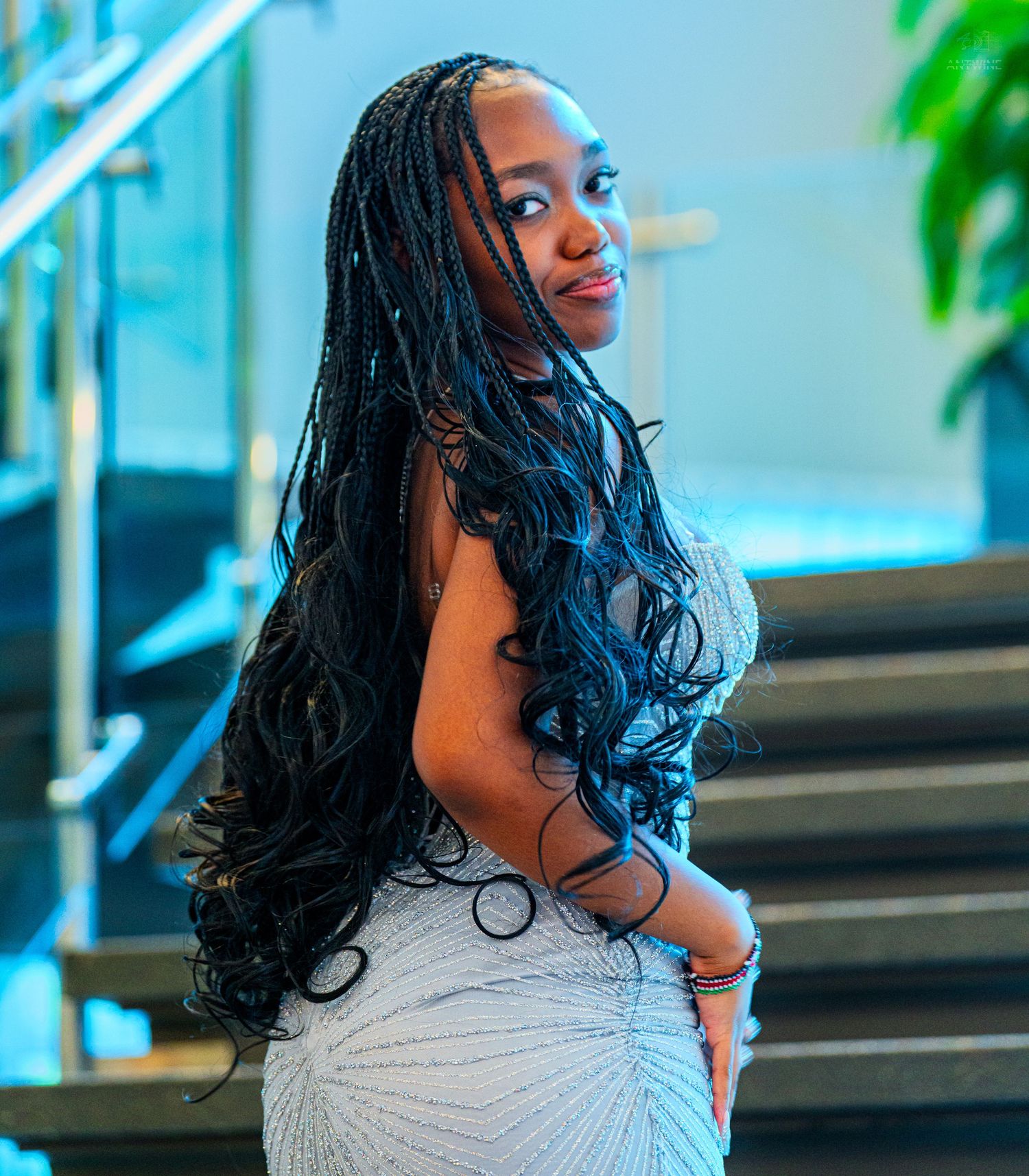 Young Black woman in white sequin dress with long braided hair poses gracefully on stairs in elegant venue.