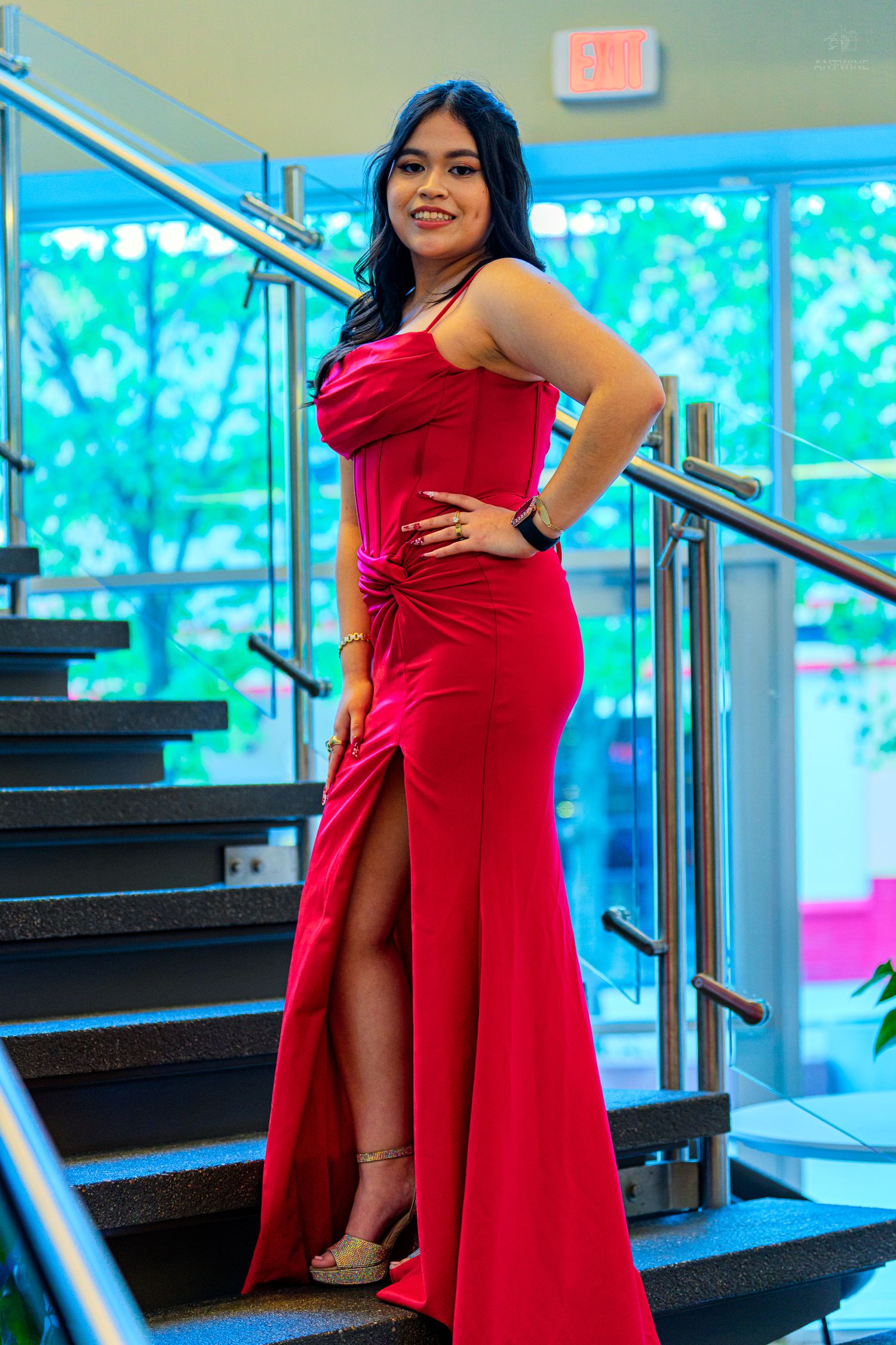 Young woman in red formal gown with slit posing on stairs at prom or formal event.