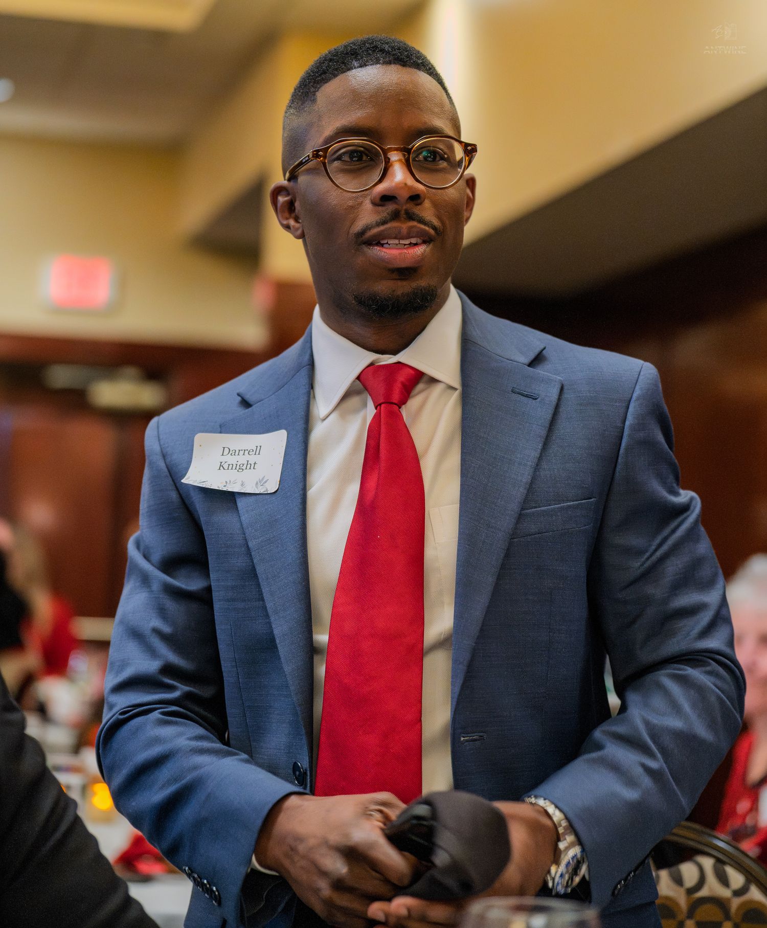 Young professional man in blue suit and red tie wearing name tag at formal networking event.
