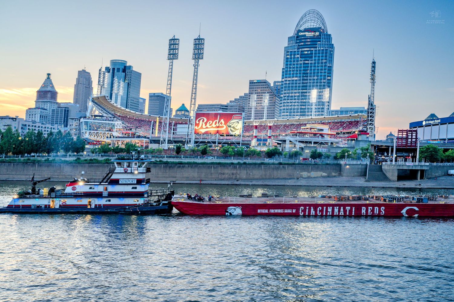 Cincinnati Reds stadium glows at sunset along the Ohio River with a barge in the foreground.